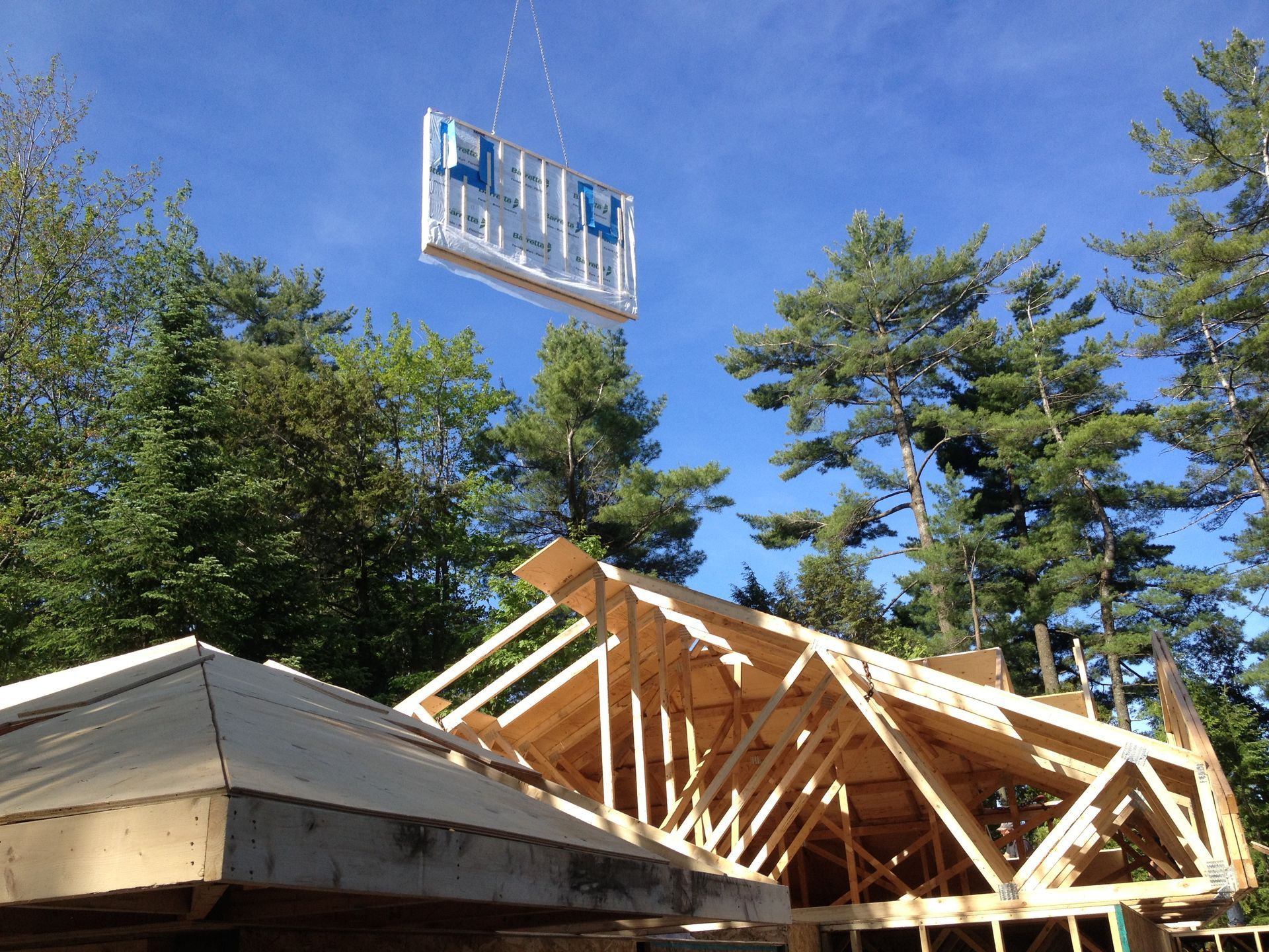 A wall panel being lifted by a crane towards a partially constructed roof frame on a sunny day.