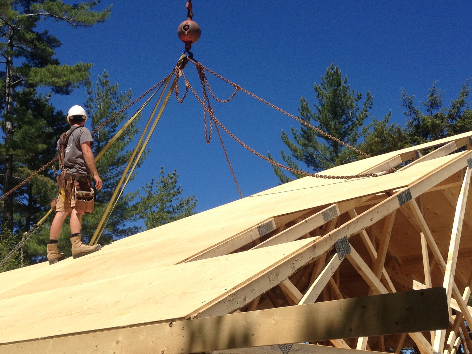 Construction worker on roof with crane lifting plywood for roof. Blue sky.