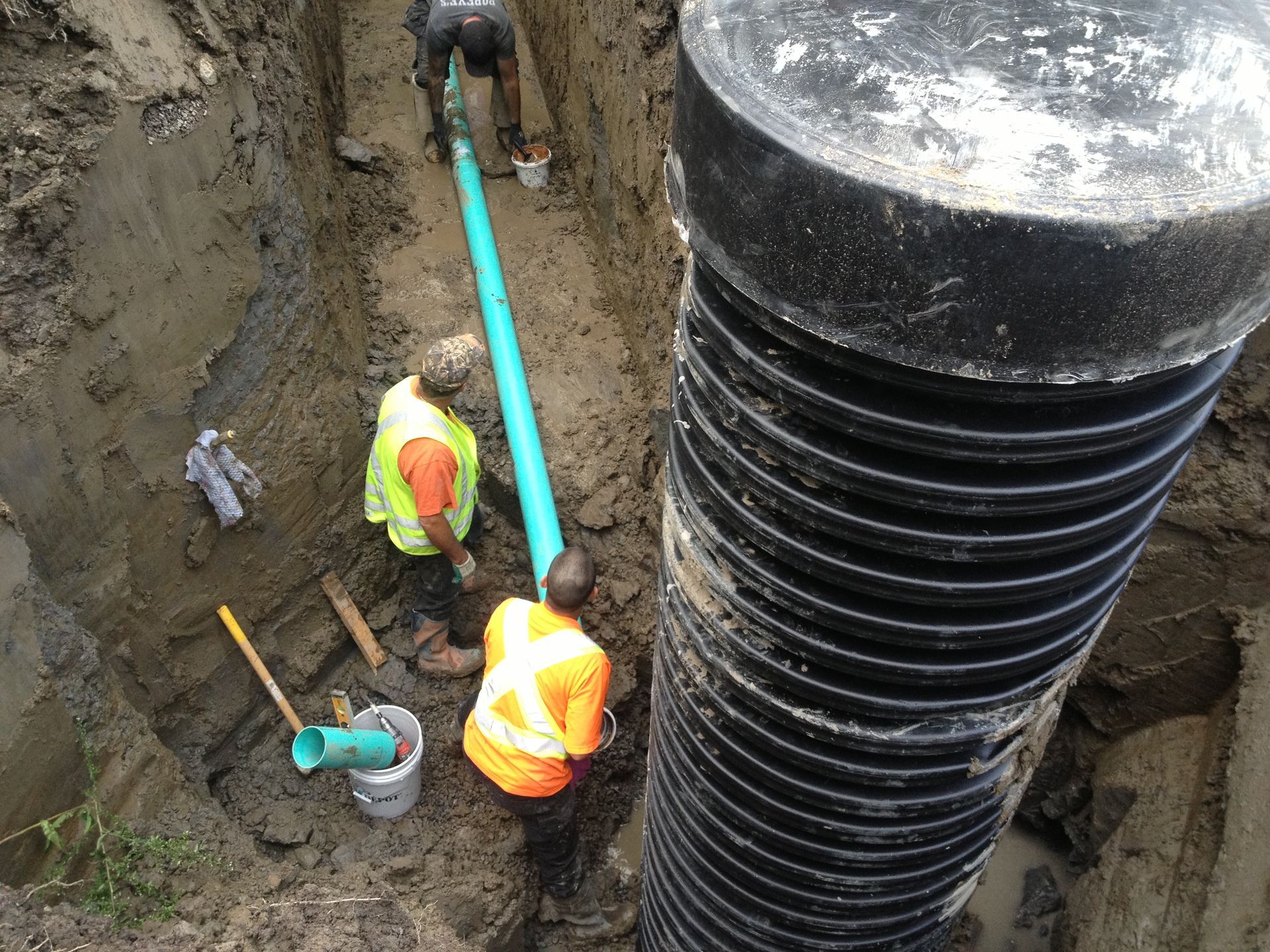 Workers installing green pipe in a trench beside a large black corrugated structure.