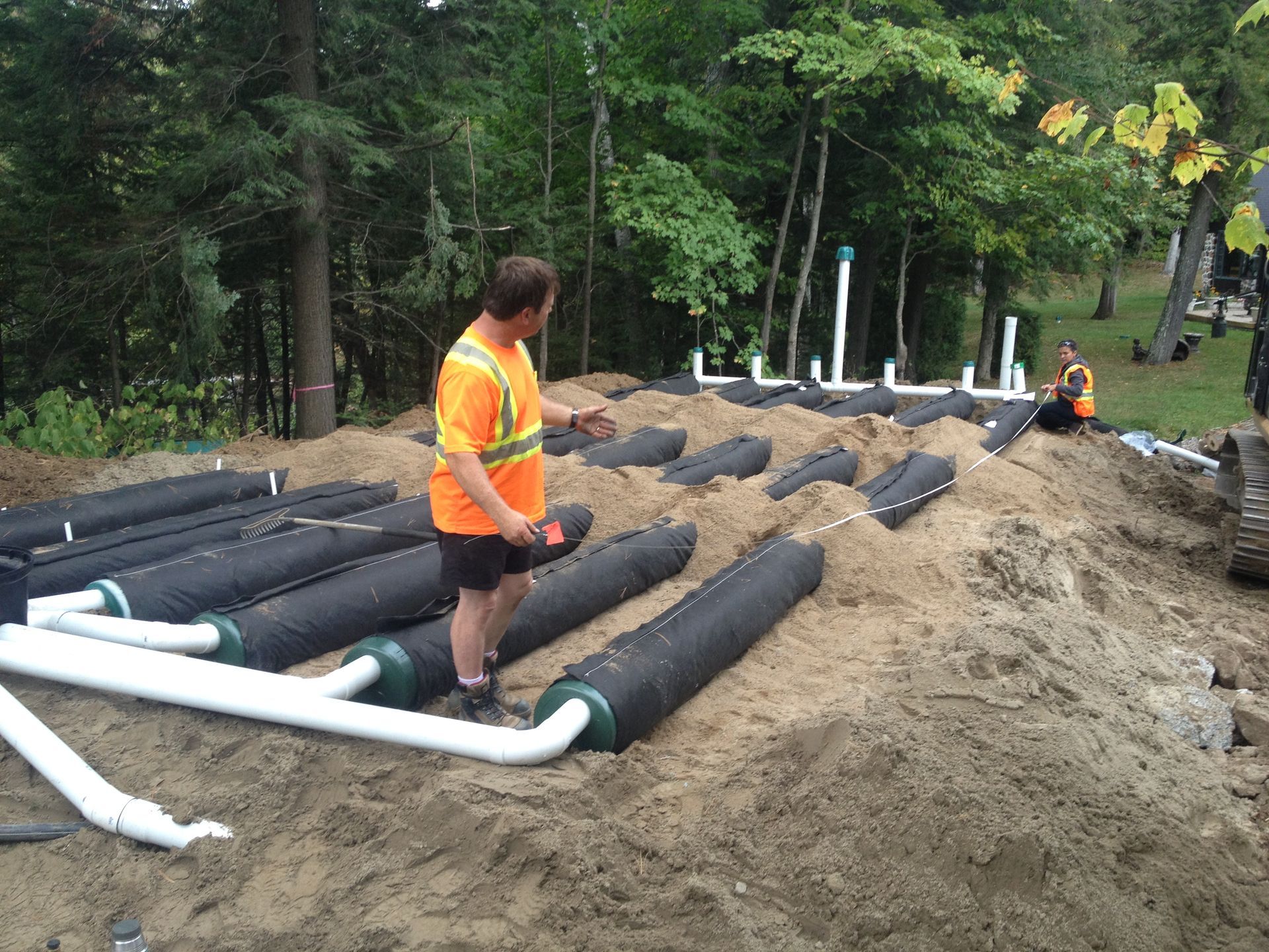 Construction workers installing a septic system. Cylindrical tanks, white pipes, and sand on a hillside.