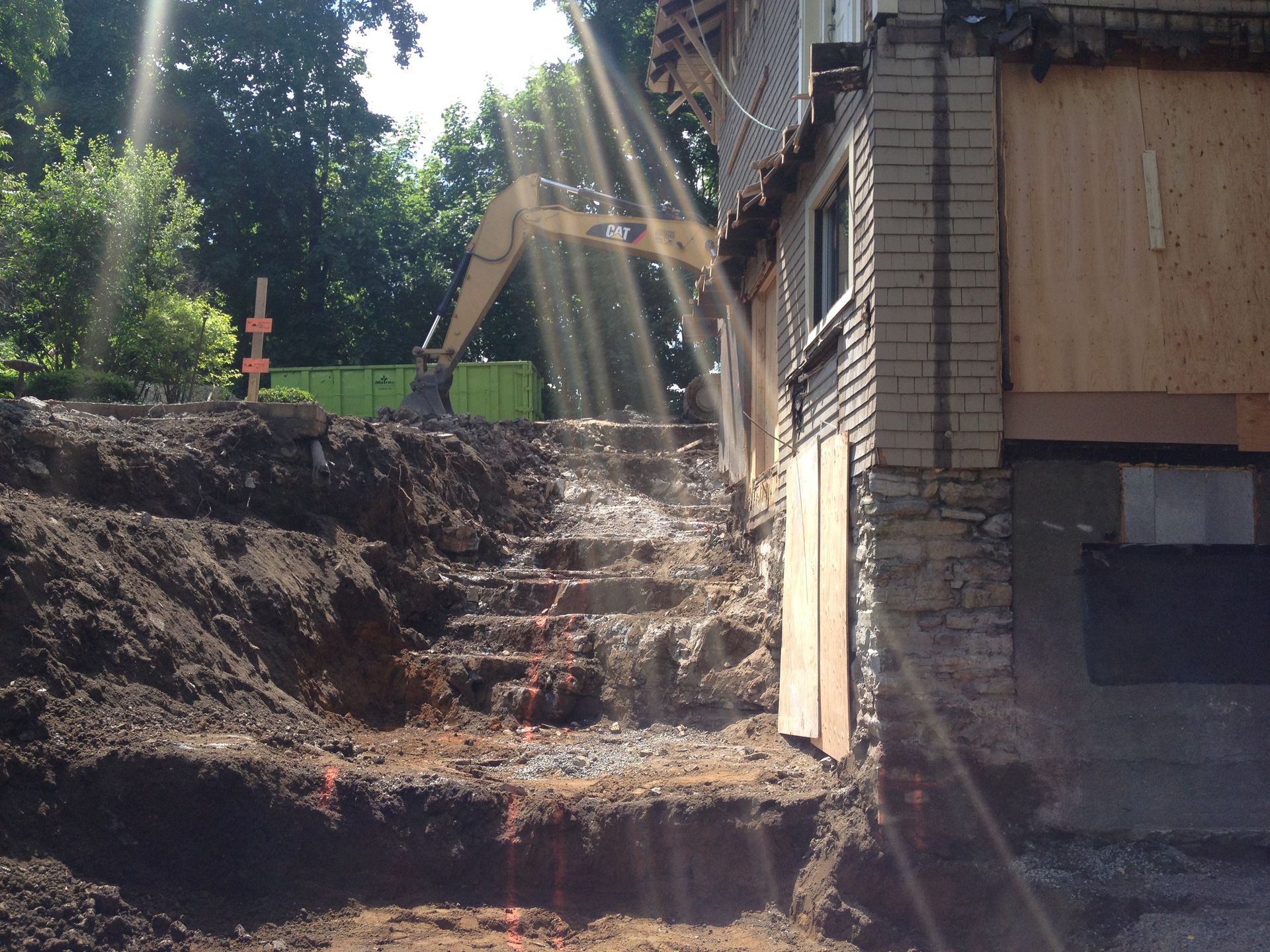 Excavation around a building with stairs, an excavator, and a debris container, outdoors in sunlight.