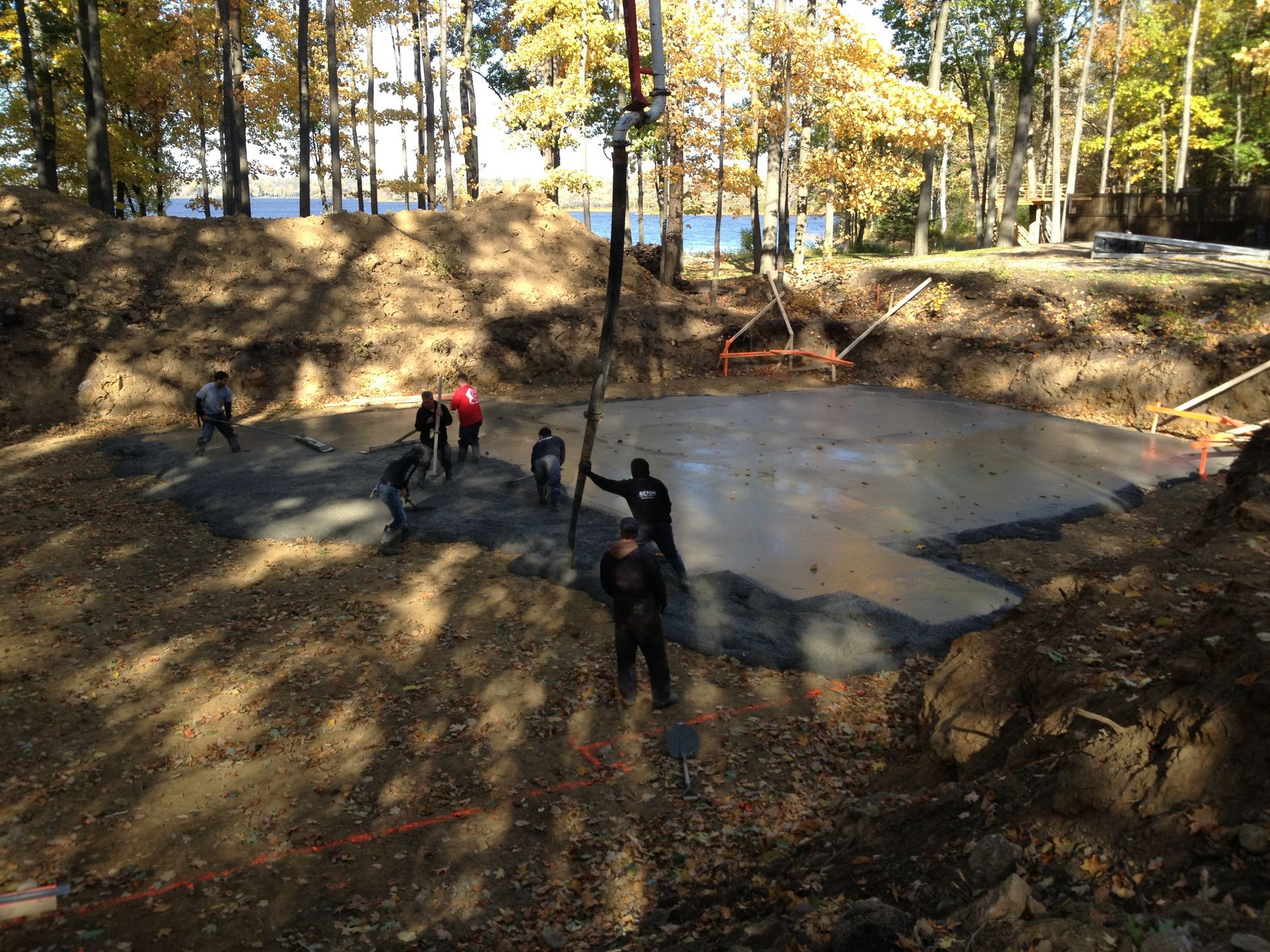 Construction crew pouring concrete into a large excavated area in a wooded setting.