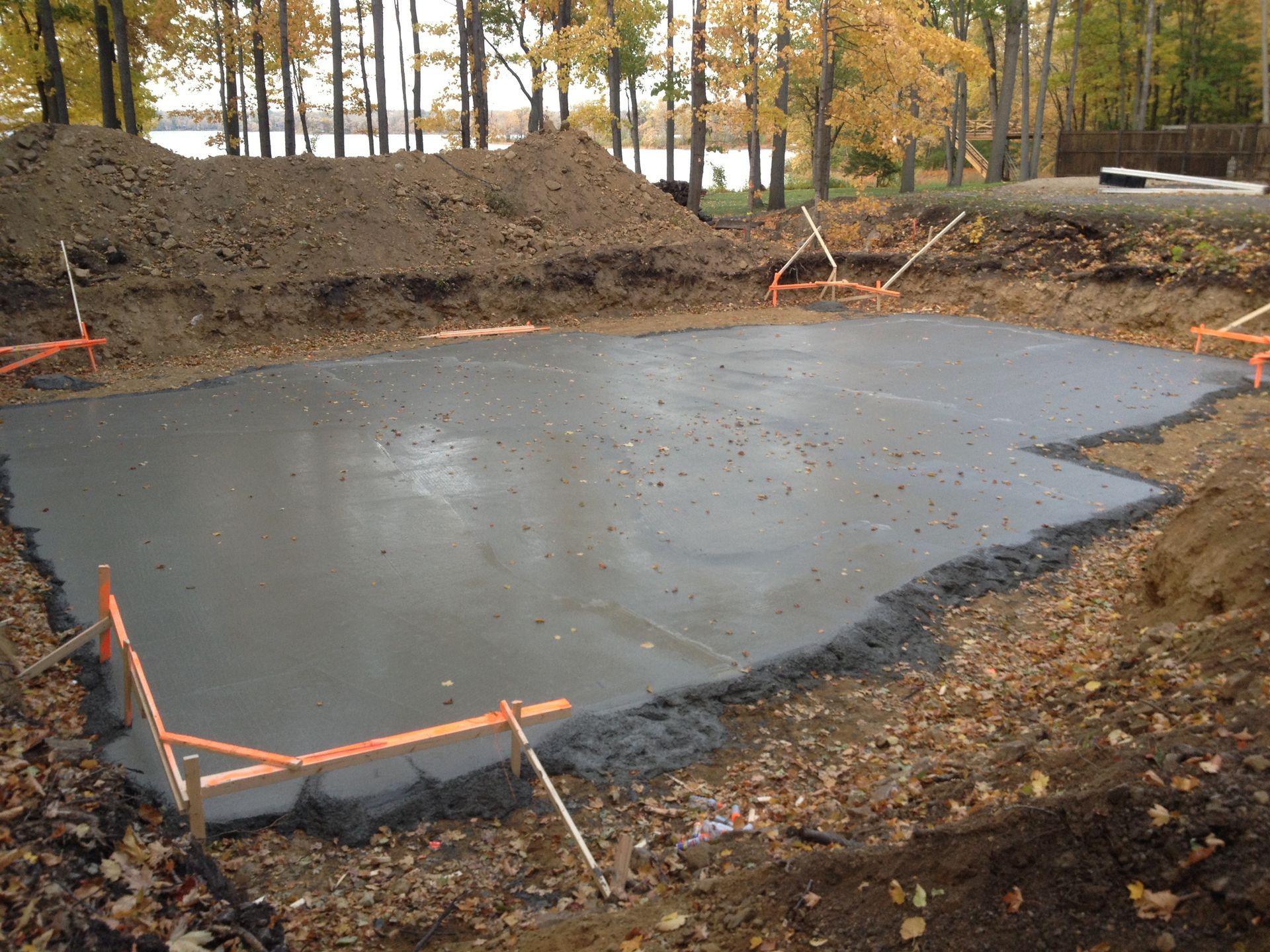 Freshly poured concrete foundation in an excavated area, with orange stakes and autumn trees in the background.