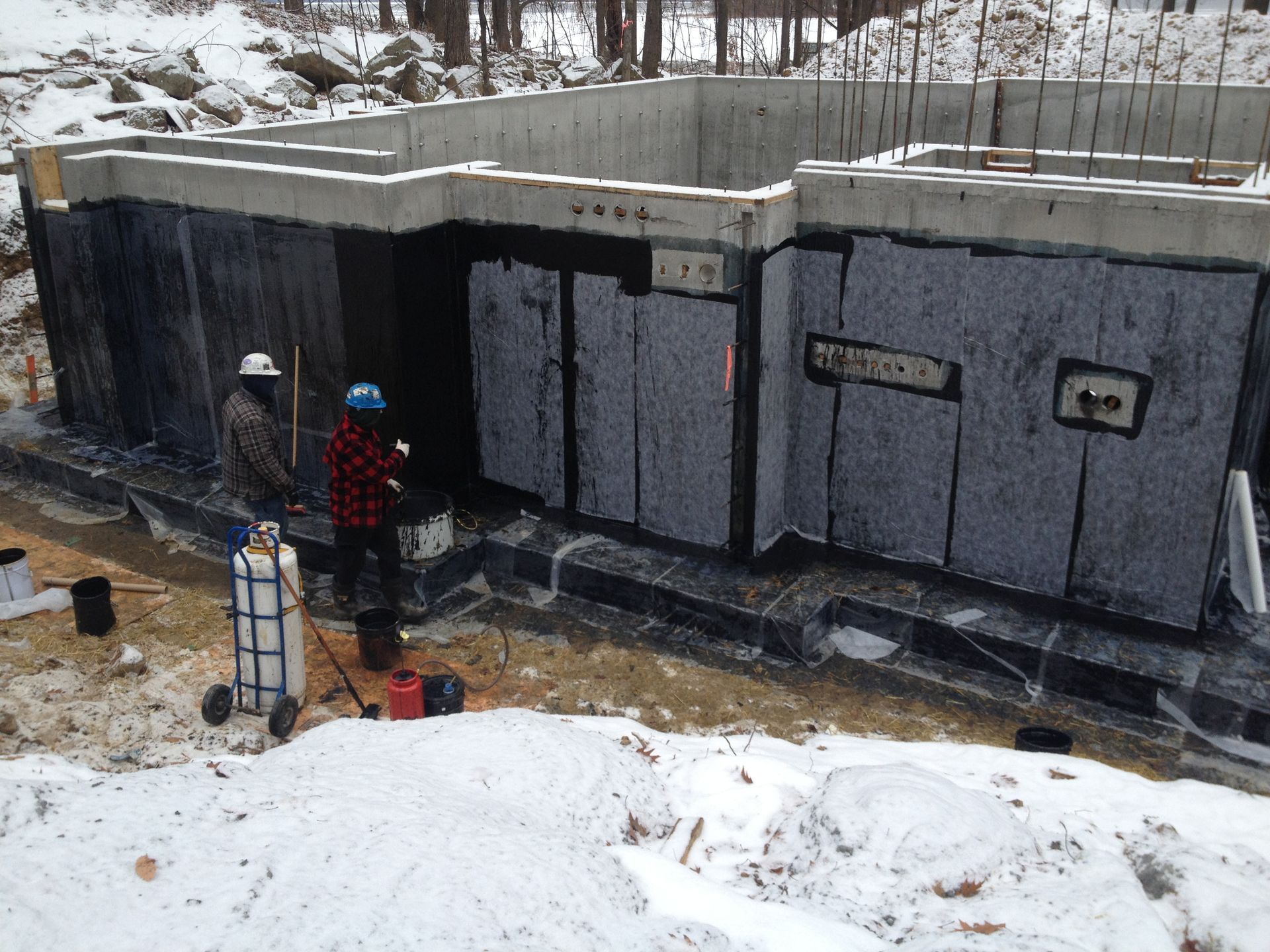 Construction workers applying insulation to a concrete foundation in a snowy setting.