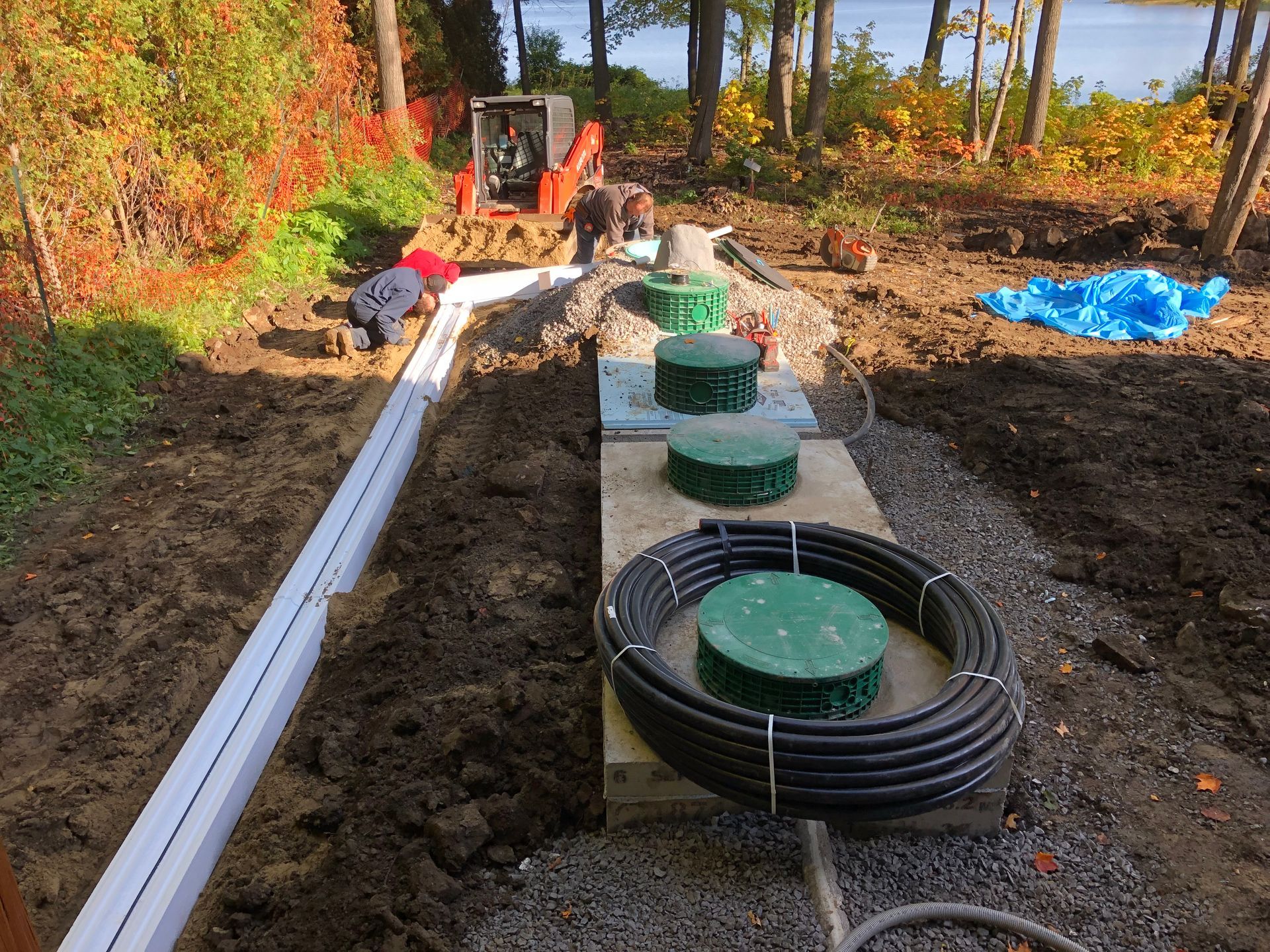 Construction of septic system: pipes, tanks, and excavator on dirt near a body of water.