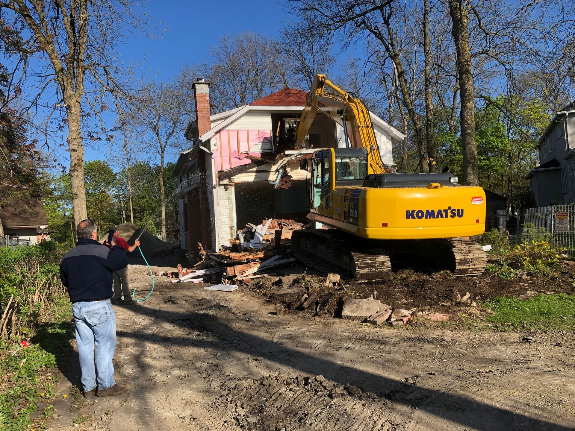 A yellow excavator demolishes a house; two people watch.