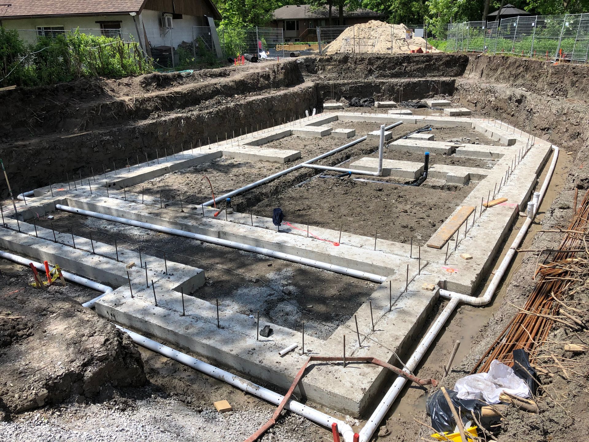 Concrete foundation under construction, with plumbing, rebar, and excavated earth surrounding it.
