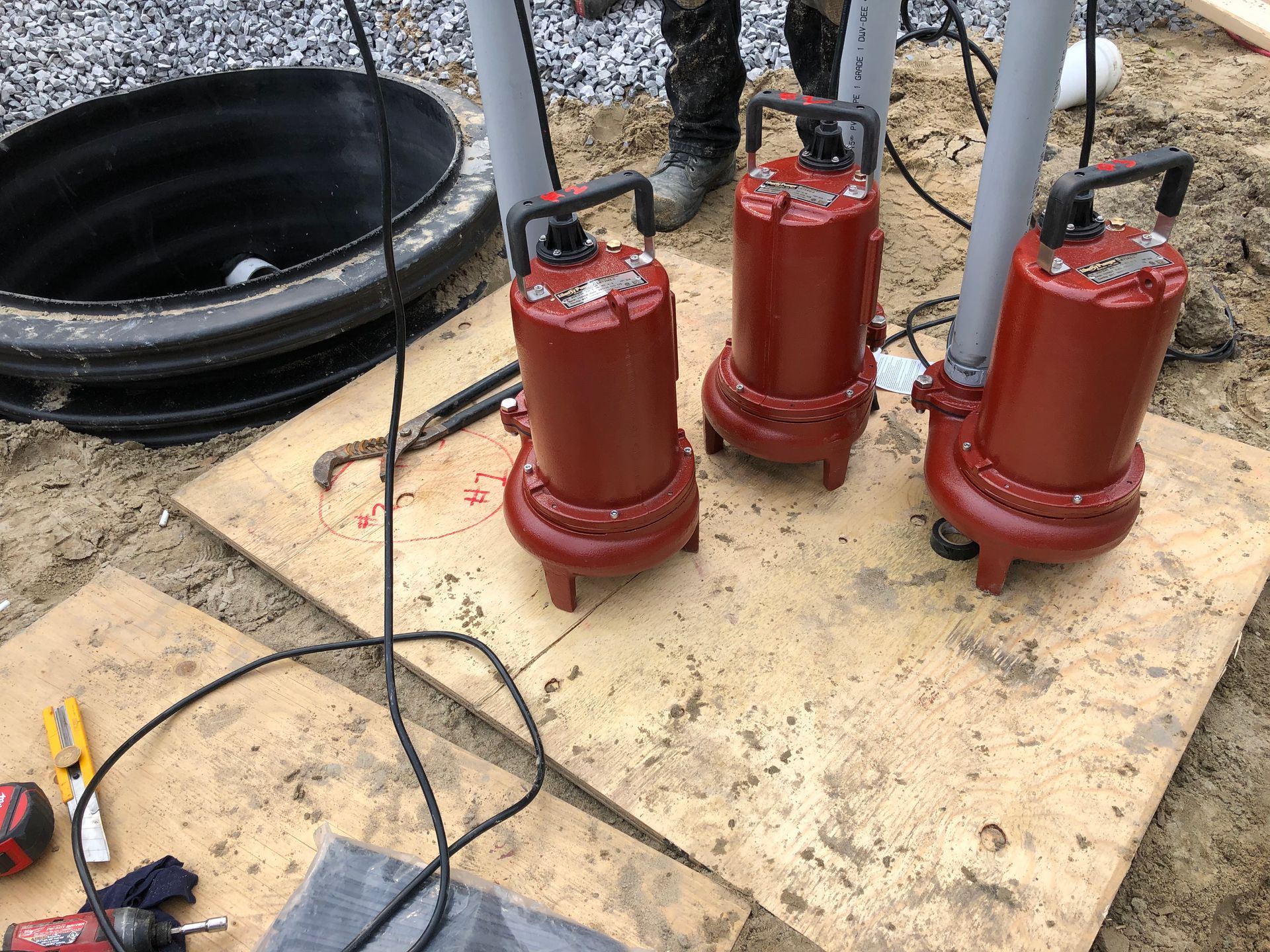 Three red submersible pumps on wooden boards next to a black well, pipes, and tools.