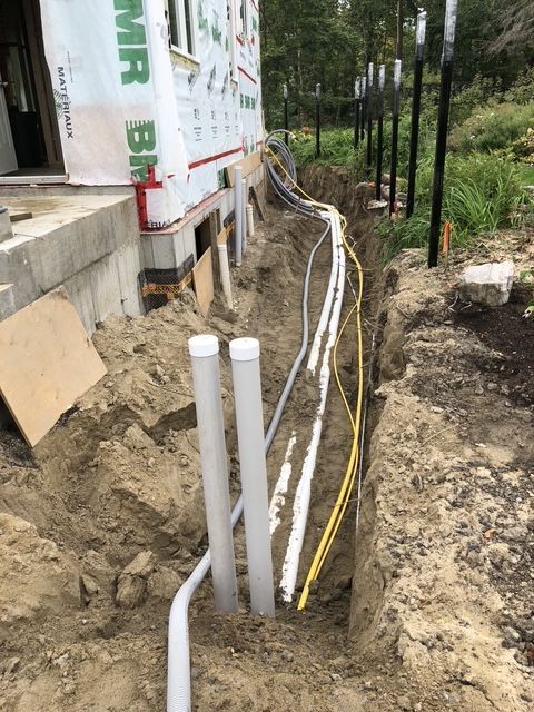 Trench alongside a building foundation holding various pipes and conduits. Dirt, gray, white, and yellow lines present.