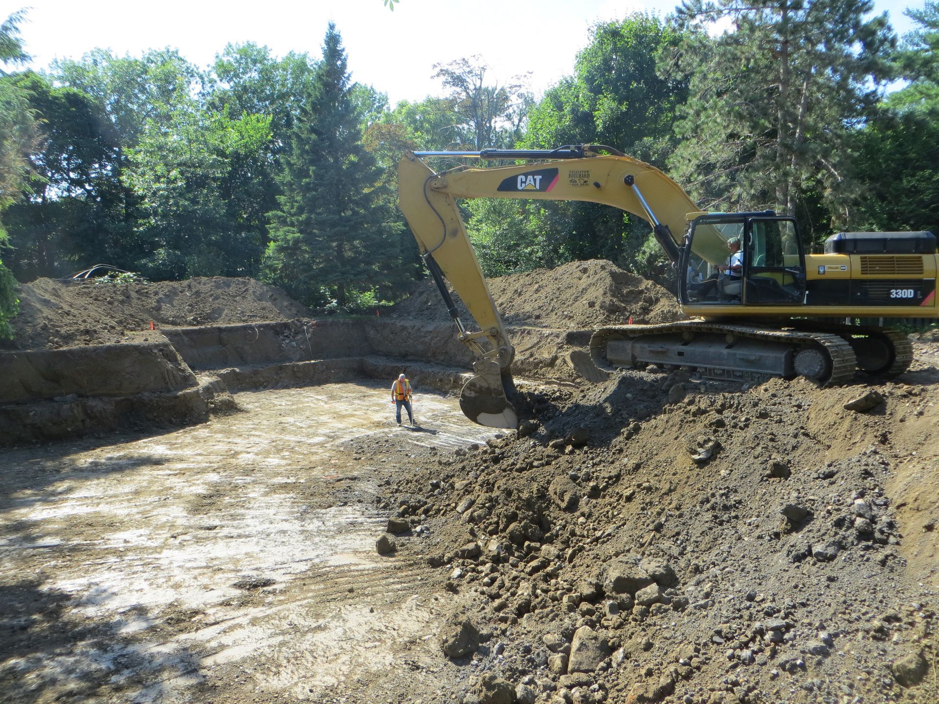 Excavator digging in an earthen pit. Man in orange vest stands in pit. Sunny day, trees in background.