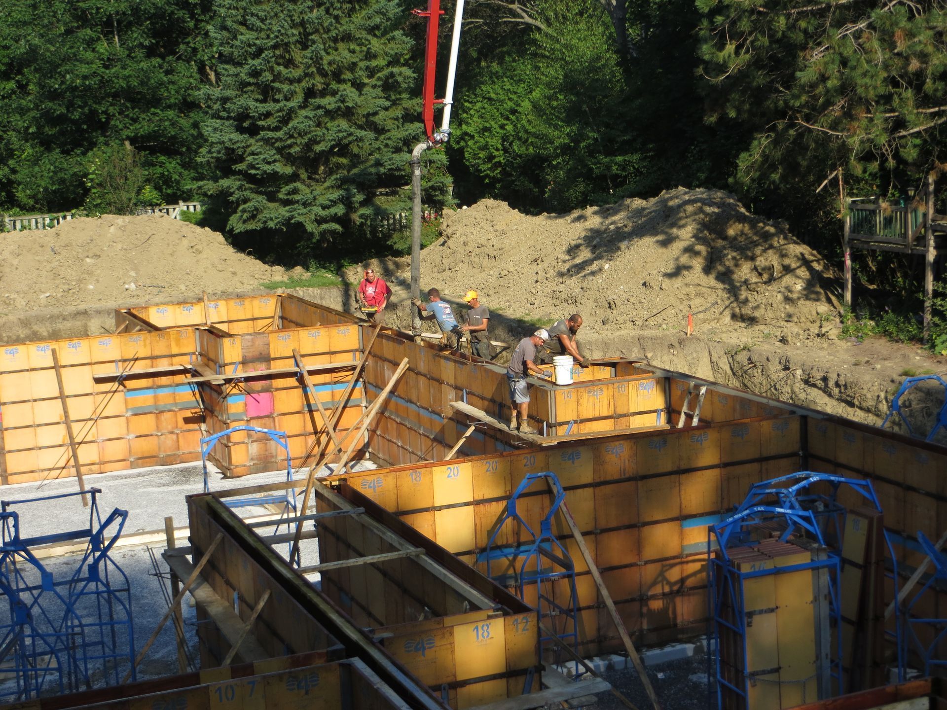 Construction workers pouring concrete into wooden forms at a building site; pump truck, trees in background.