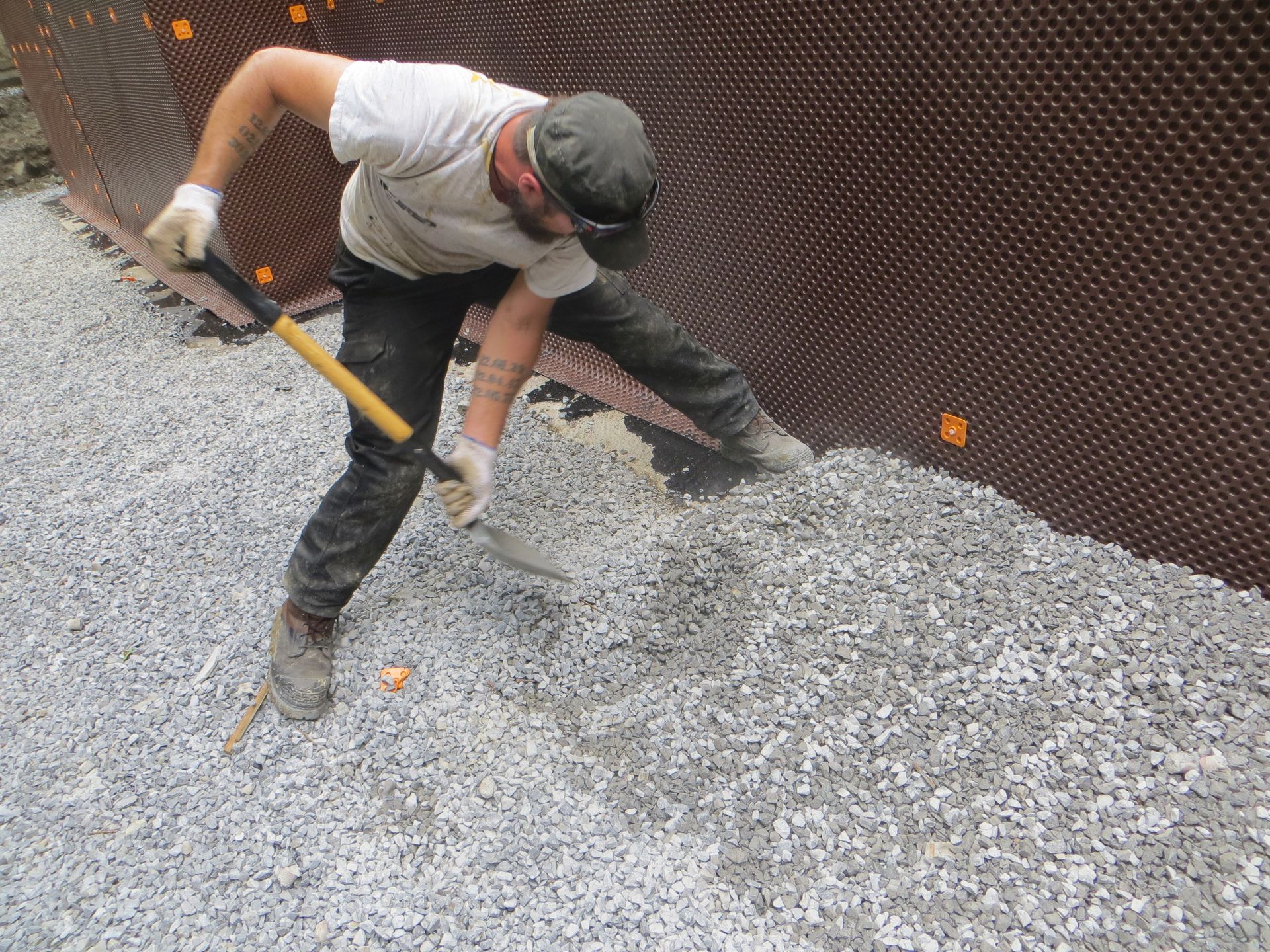 Construction worker shoveling gravel next to a dark brown foundation wall with dimple membrane.