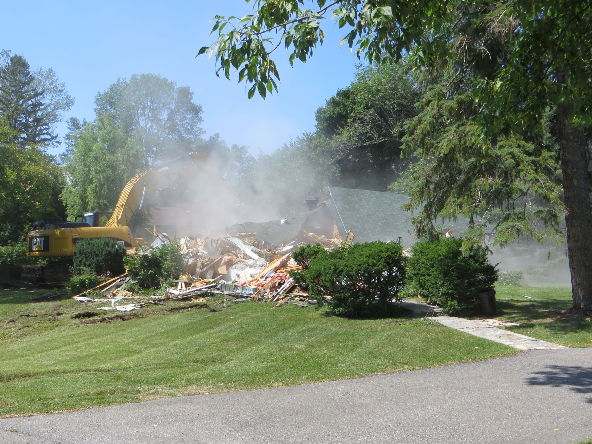 A yellow excavator demolishes a house, kicking up dust; green lawn and trees surround.