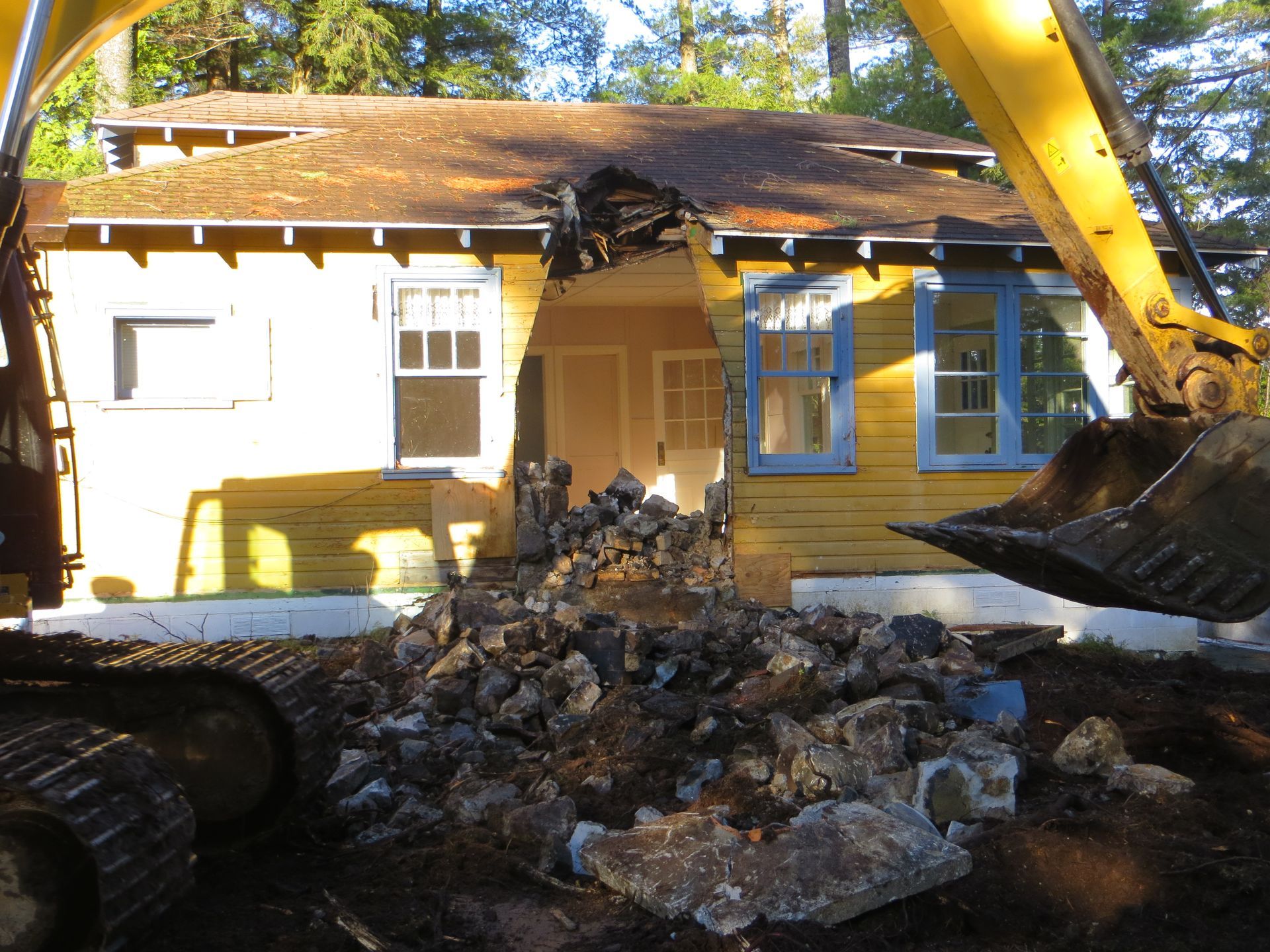 Excavator demolishing a yellow house, debris spilling out.