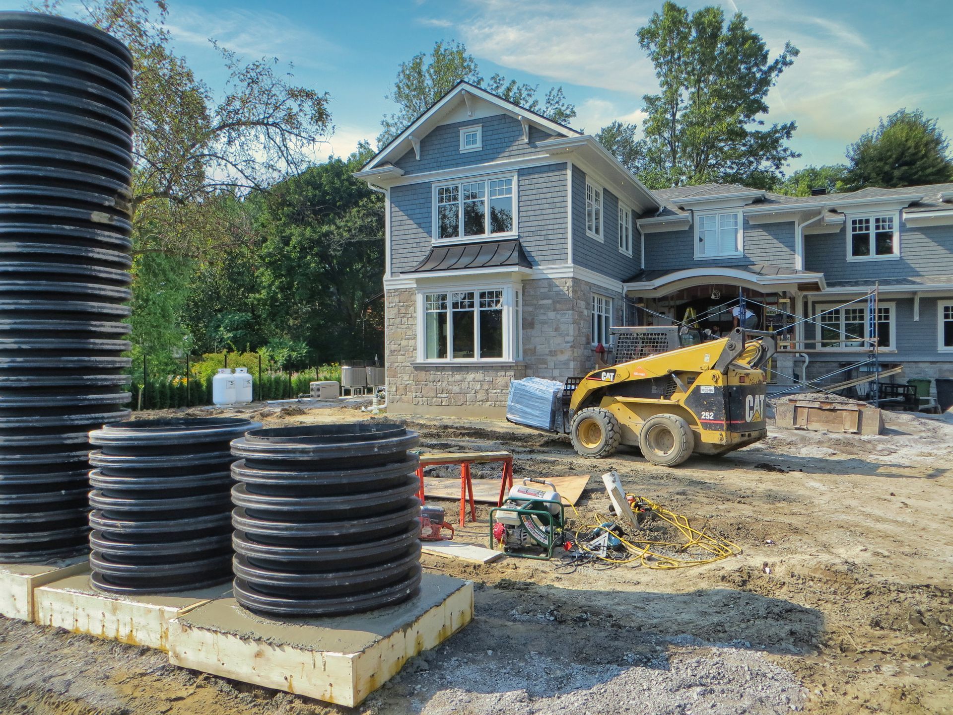 Construction site with a two-story house, a skid steer, and corrugated plastic pipes.