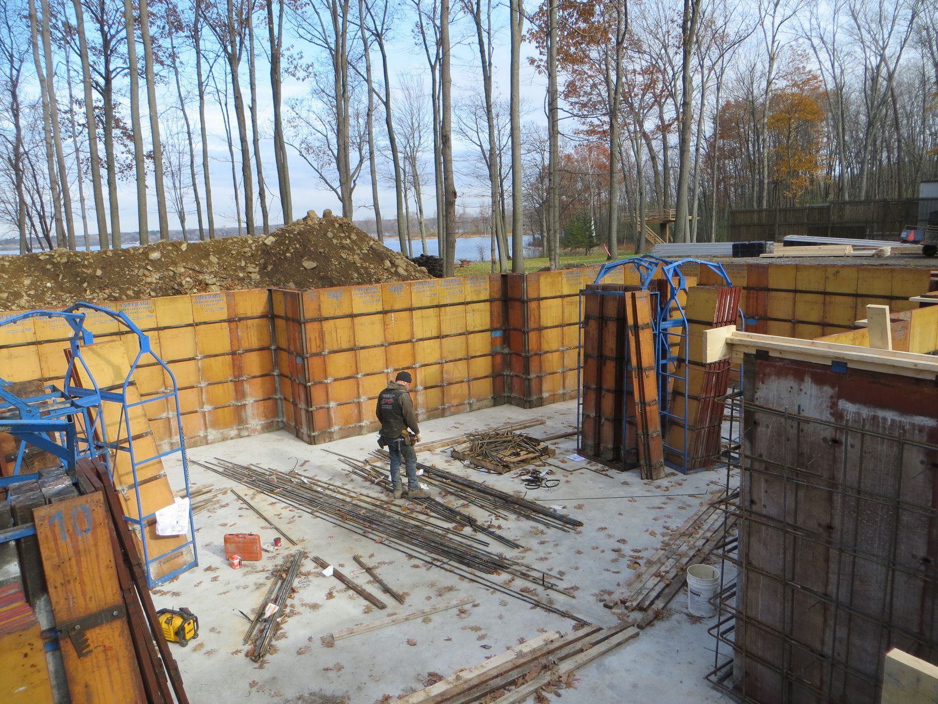 Construction site: concrete forms, rebar, worker, trees in background.