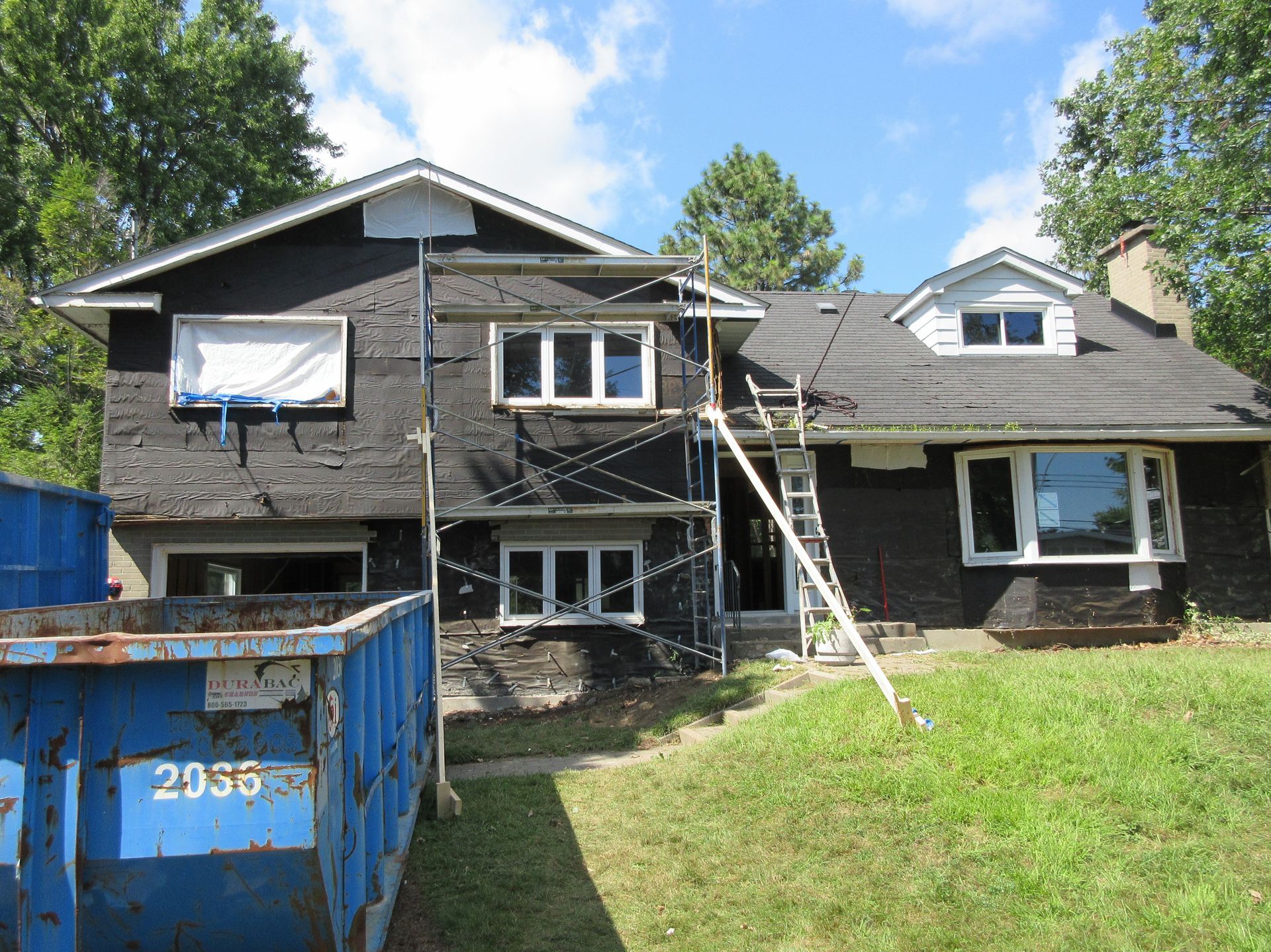 House exterior under renovation with scaffolding, blue dumpster on lawn.