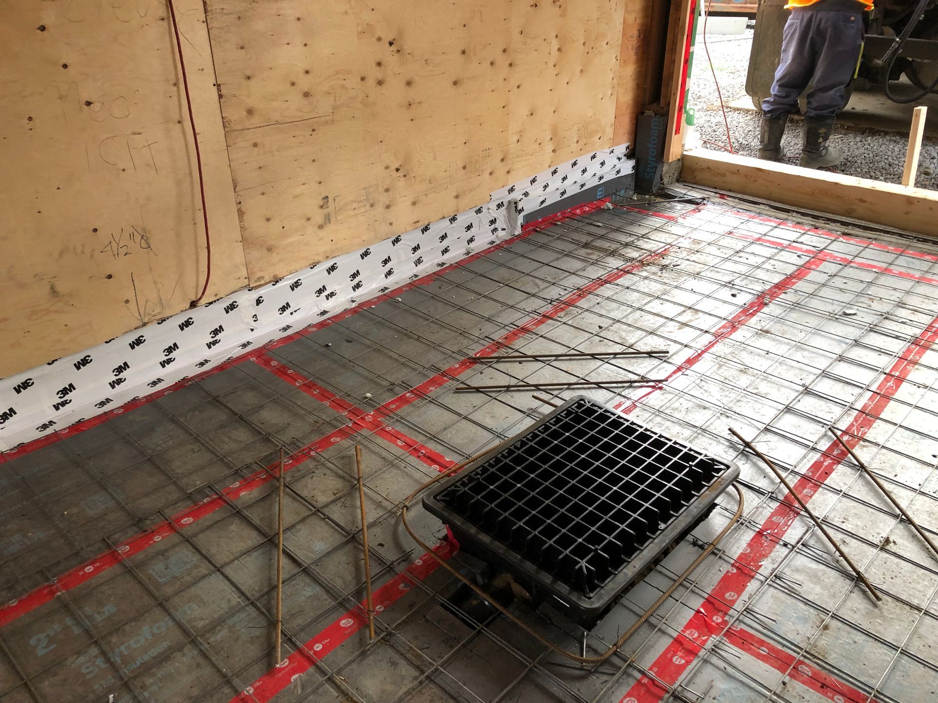 Concrete floor with red radiant heat tubing, steel grid, drain, and wall insulation. Construction site.