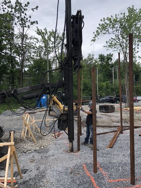 Construction site with a drilling machine and worker placing metal posts. Gravel ground and trees in the background.