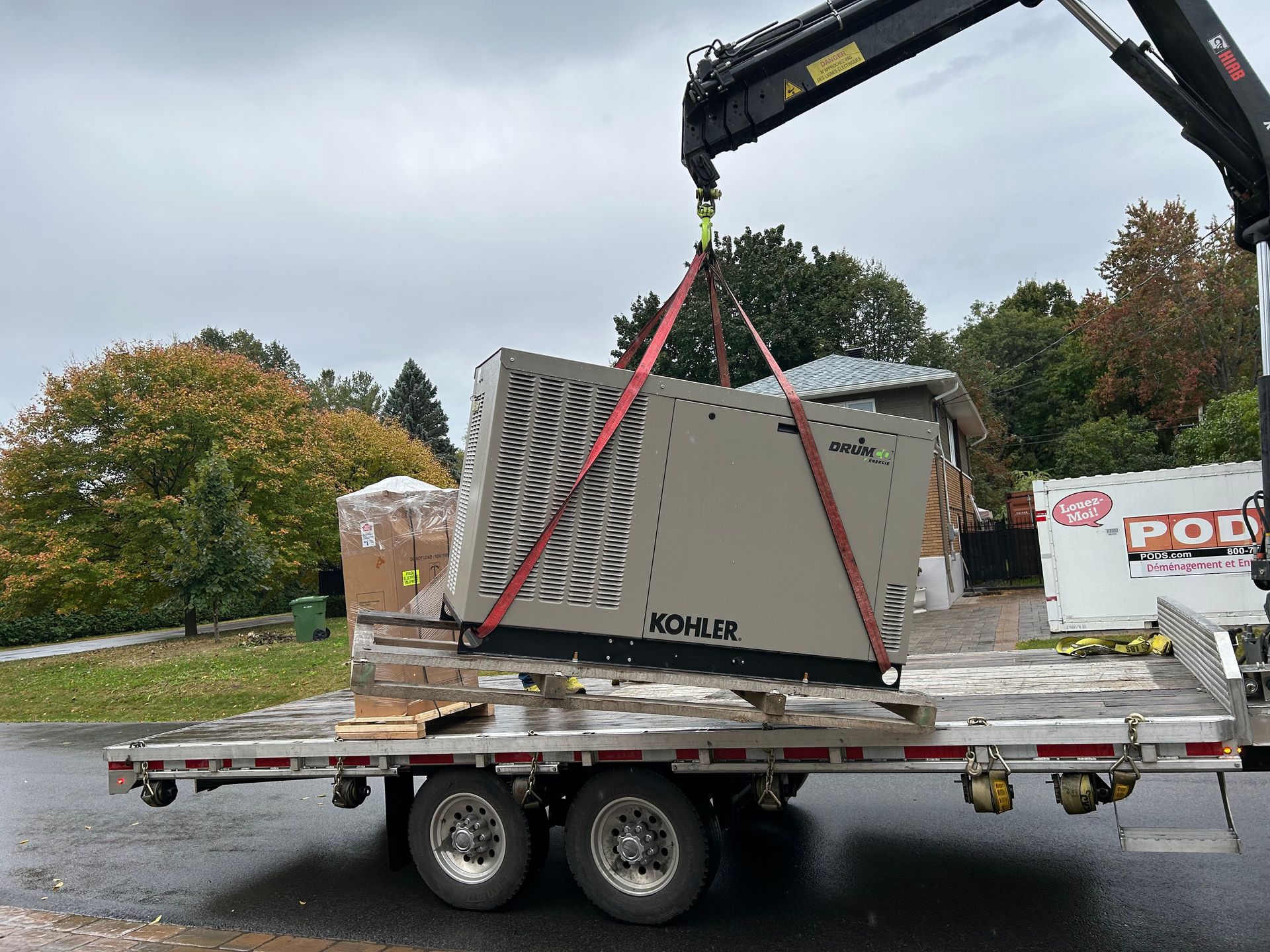 Crane lifting a Kohler generator from a flatbed trailer on a residential street. Red straps secure the generator.
