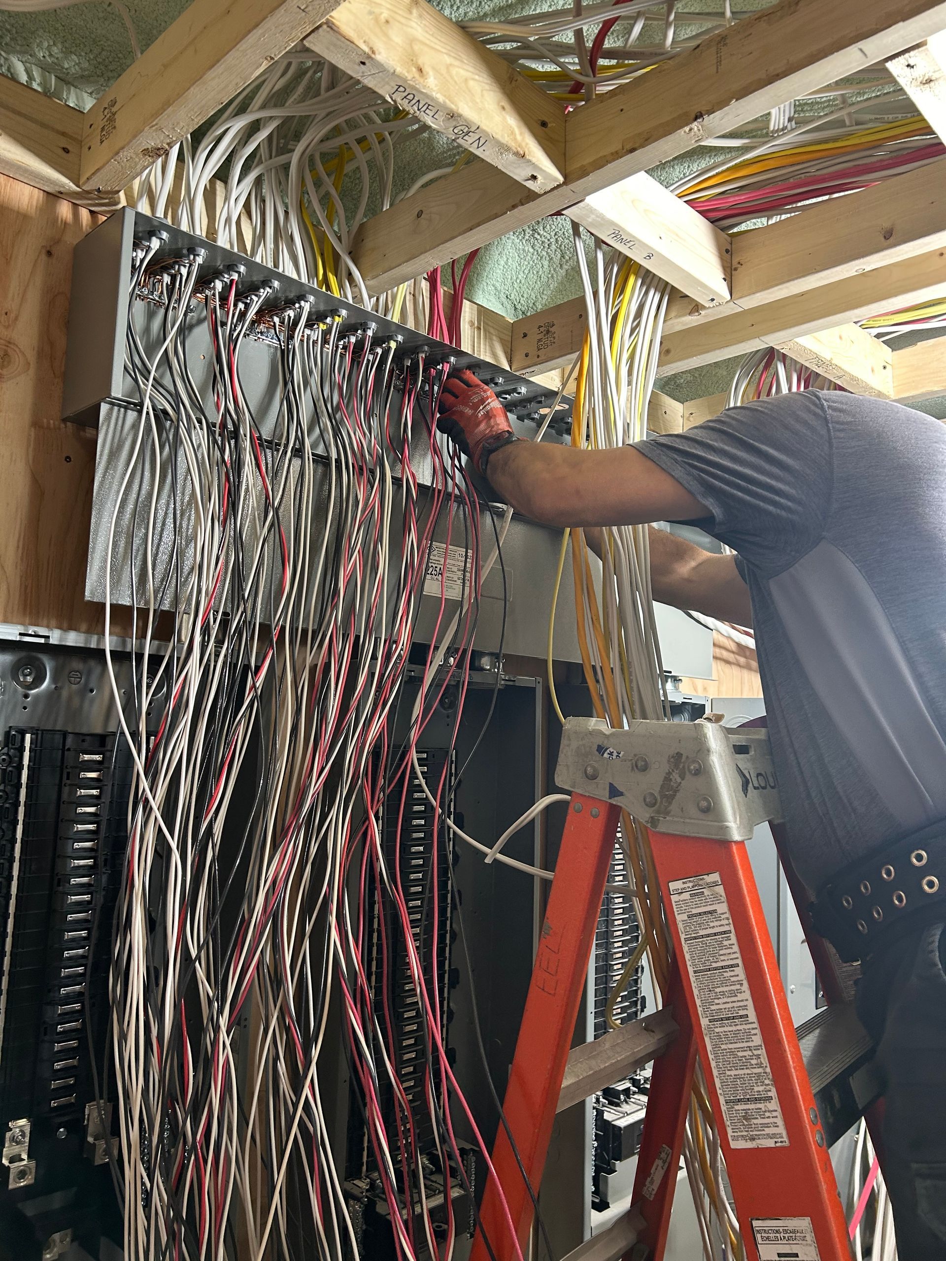 Electrician working on wiring inside an electrical panel. Red ladder, many wires, indoor setting.