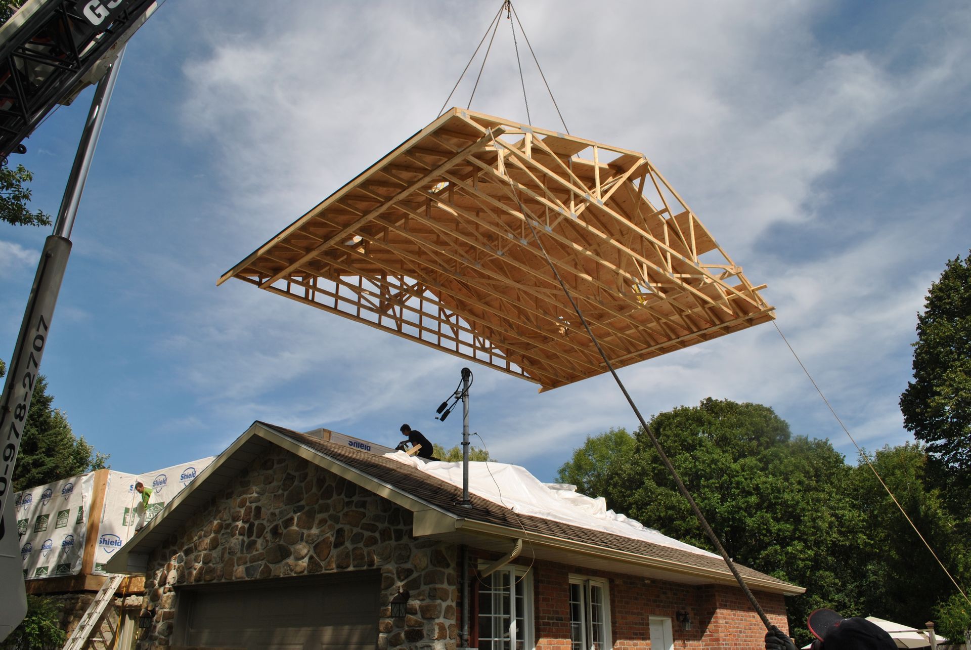 A crane lifting a large wooden roof structure over a brick building. Two workers on existing roof.