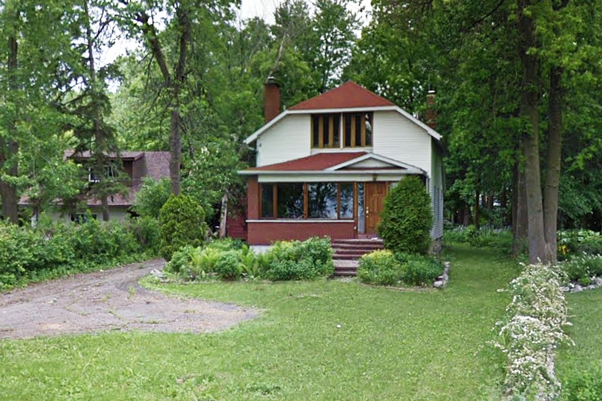 Two-story house with red roof and trim, beige siding, surrounded by trees and green grass.