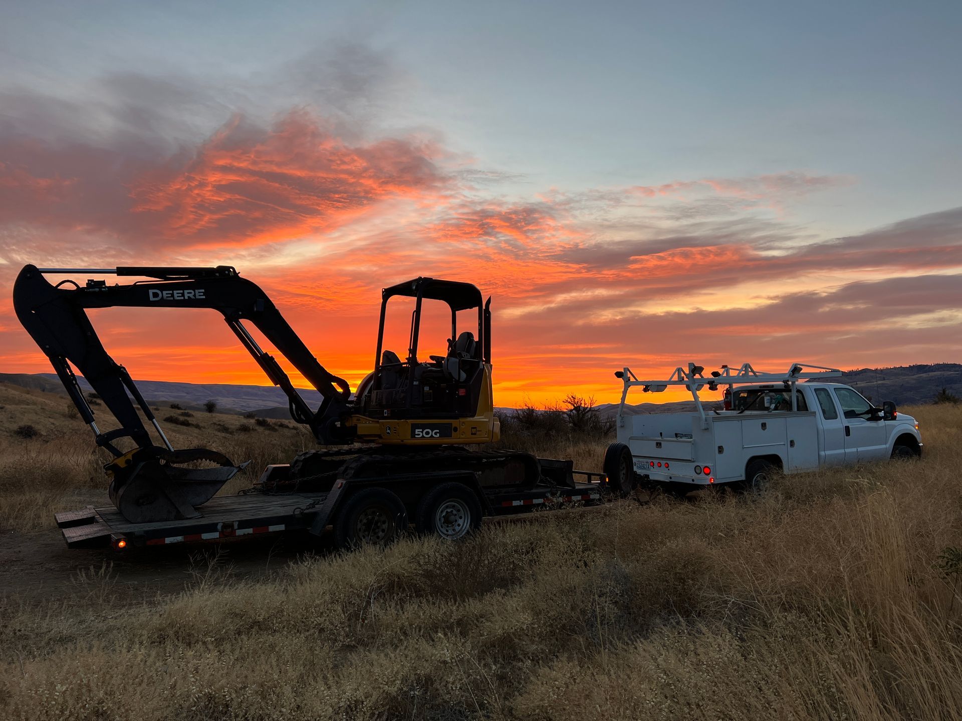 An excavator is being towed by a truck in a field at sunset.