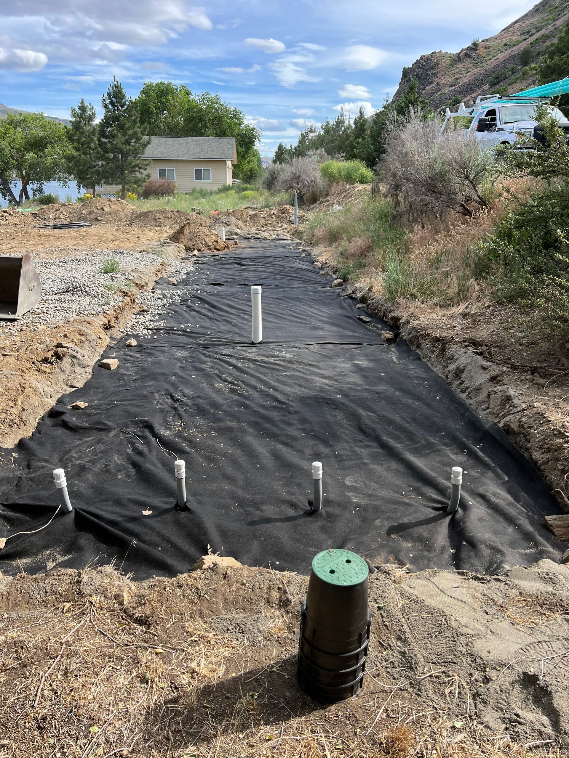 A drainage system is being built in a dirt field with a house in the background.