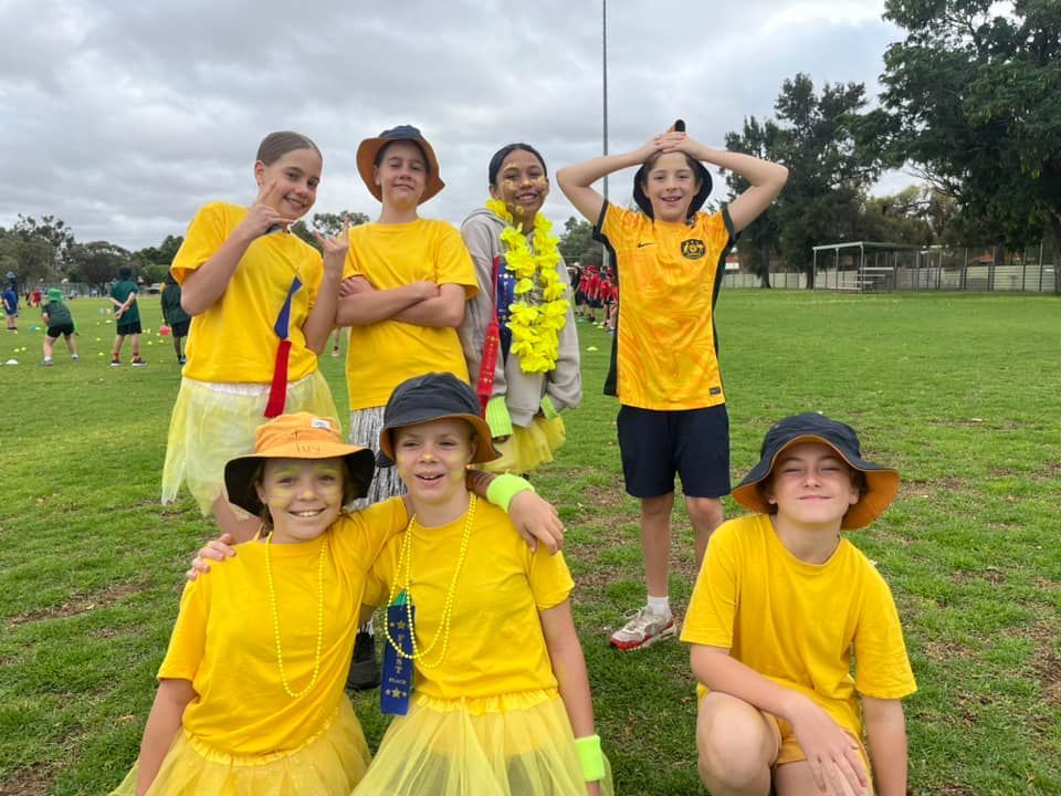 Group of children in yellow outfits pose on a grassy field, some wearing hats, leis, and tutus.
