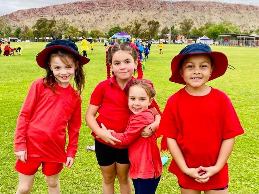 Four young children in red shirts and hats smile on a green field with a red rock formation in the background.