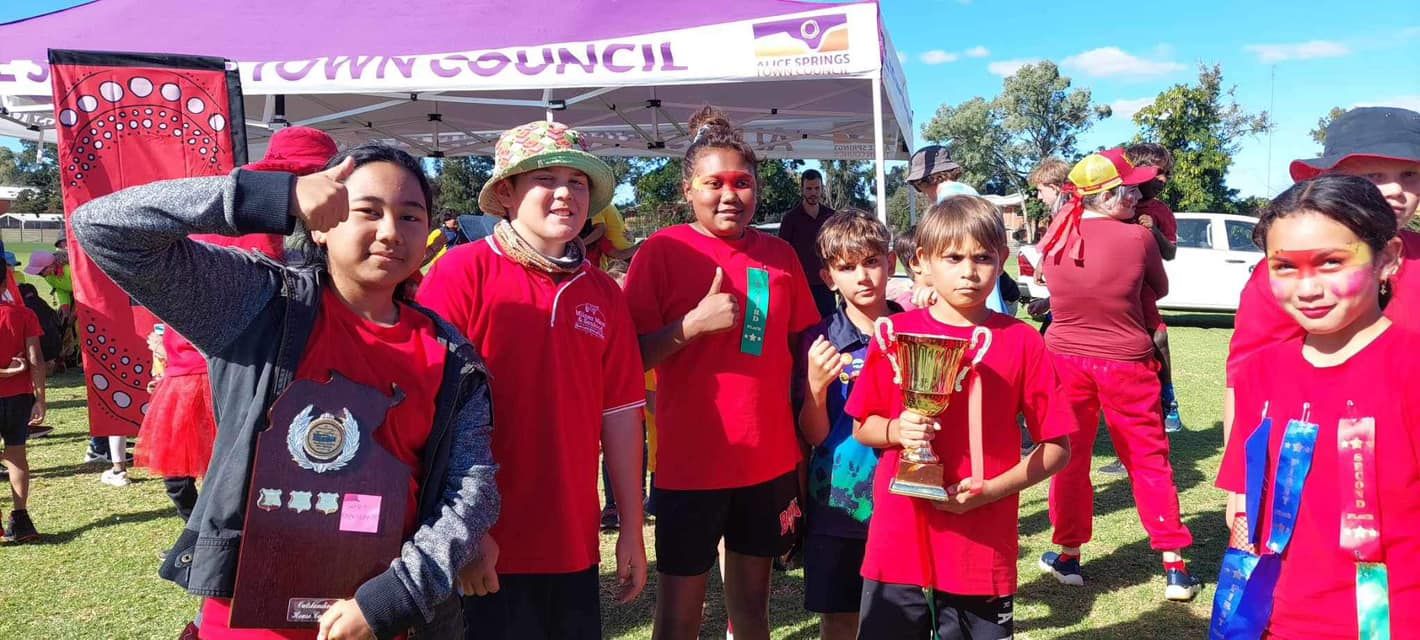 Group of children in red shirts smiling for a photo, some holding awards and wearing face paint. Outdoors.