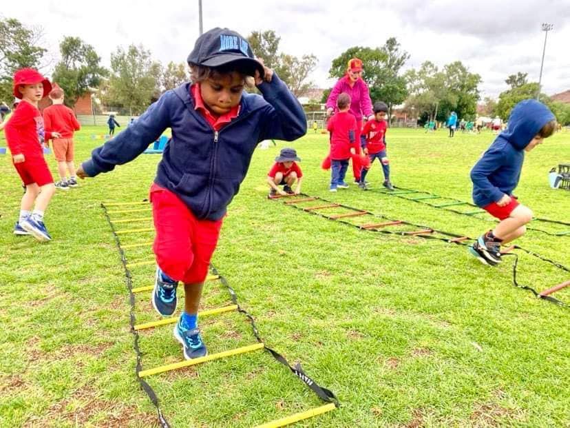 Children in red and blue outfits playing agility ladder game on a grassy field.