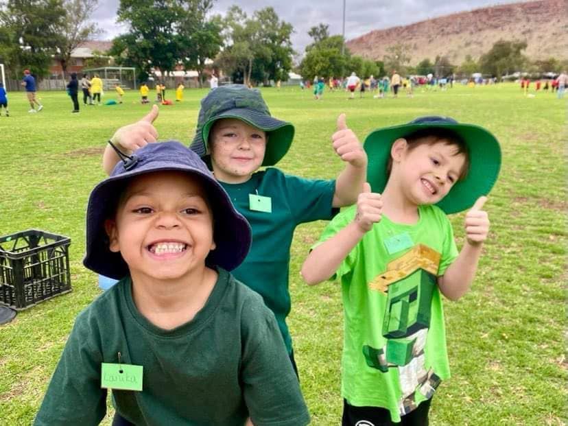 Three young children in green and blue hats giving thumbs up on a grassy field, smiling.
