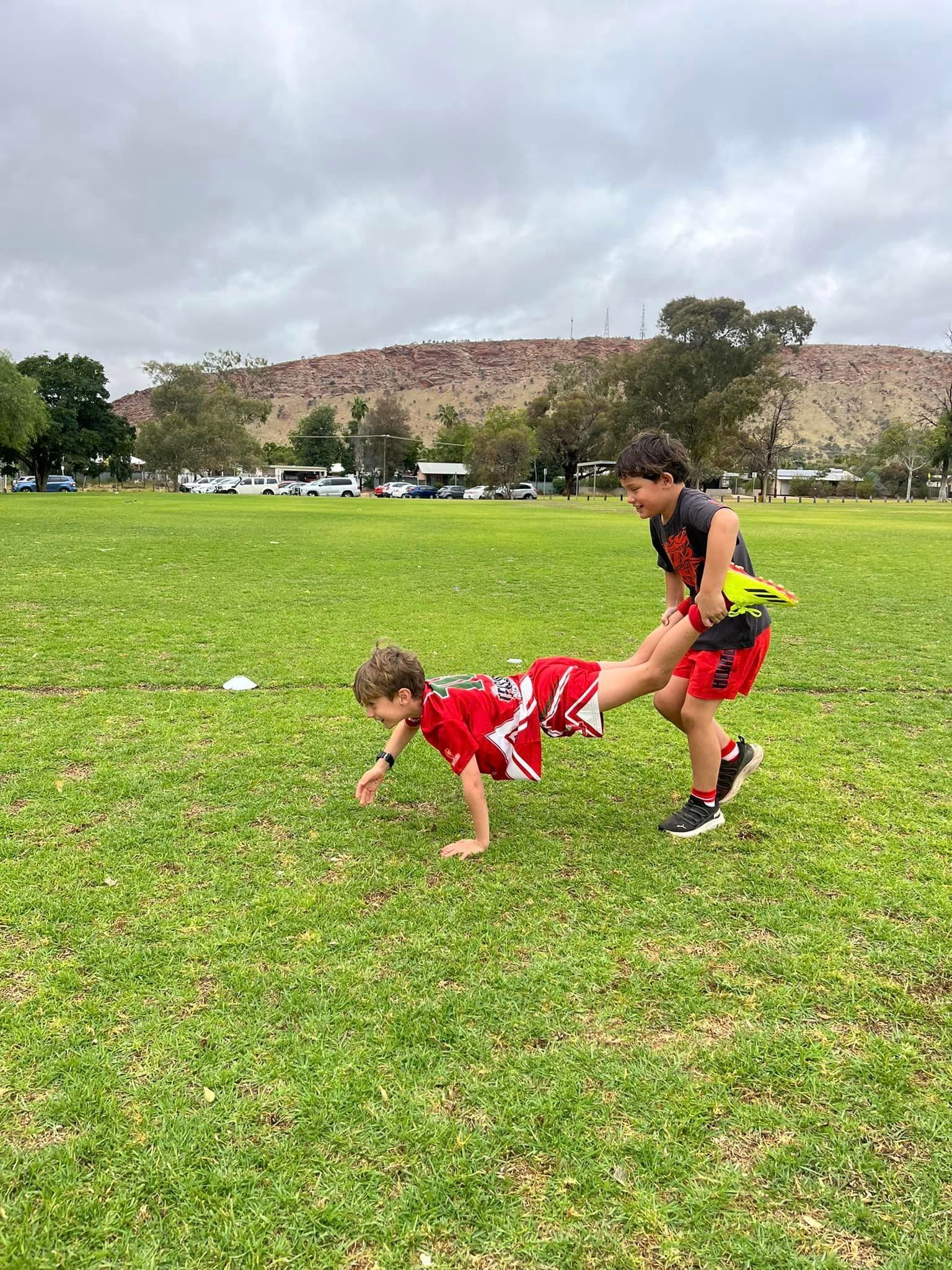 Two boys playing a wheelbarrow race on a grassy field.