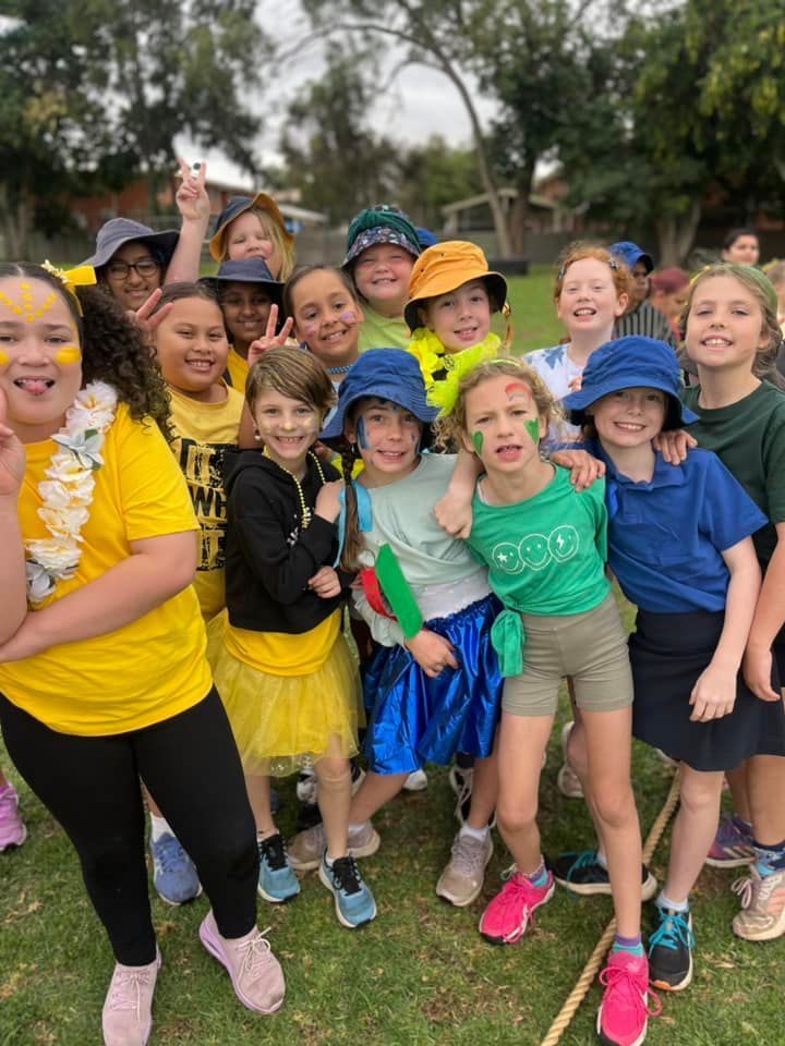 Group of children in colorful outfits, posing outdoors. Many are smiling, some with face paint, on a grassy area.