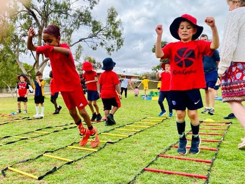 Children in red shirts and shorts jumping in agility ladders on a grass field.