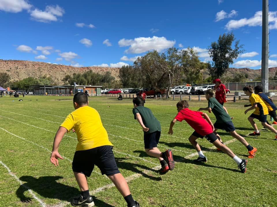 Children in a track race on a green field, with a mountain backdrop, under a blue sky.