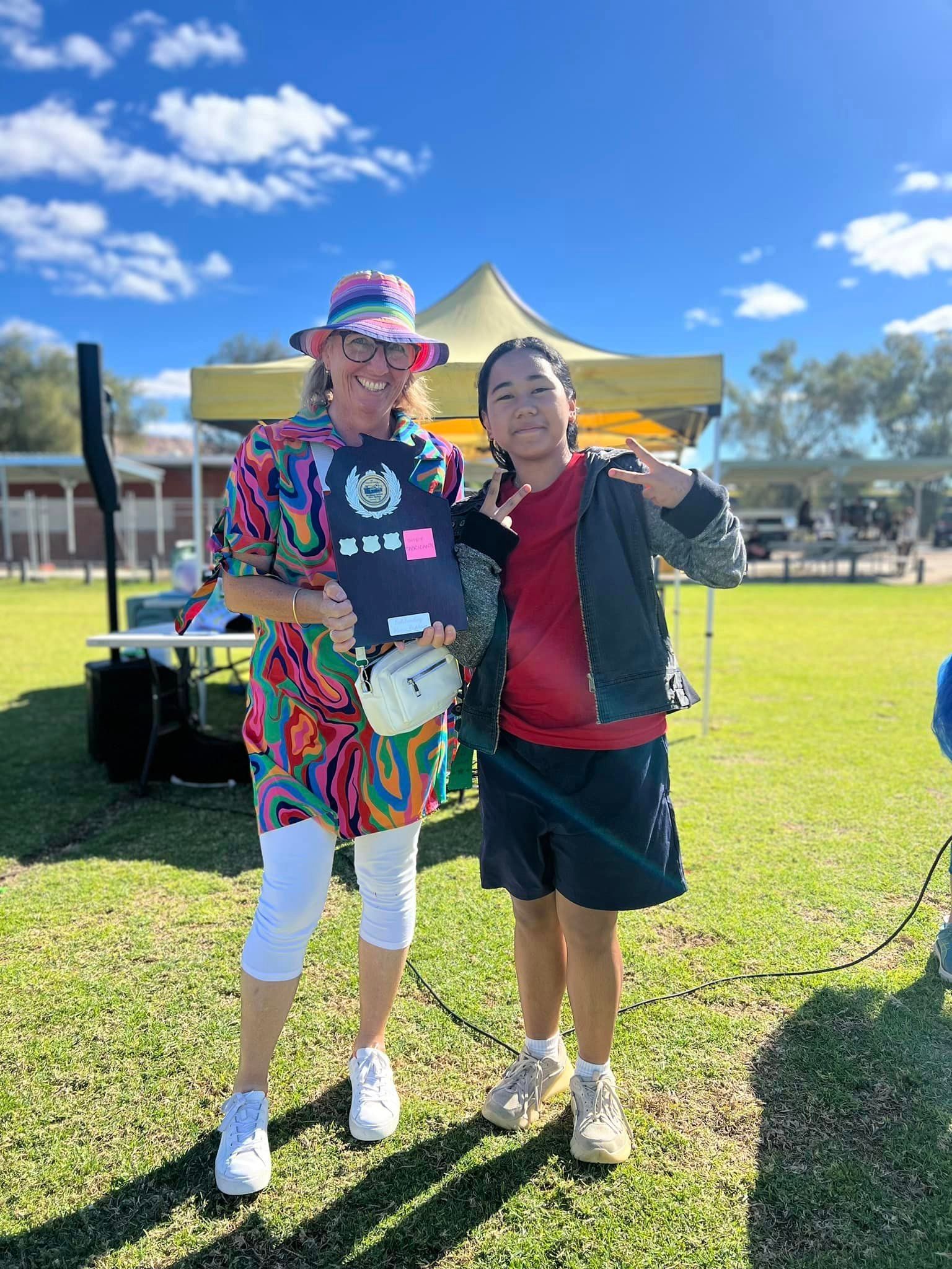 Two smiling women pose outdoors; one in colorful outfit, holding sign; the other in red top with dark jacket.