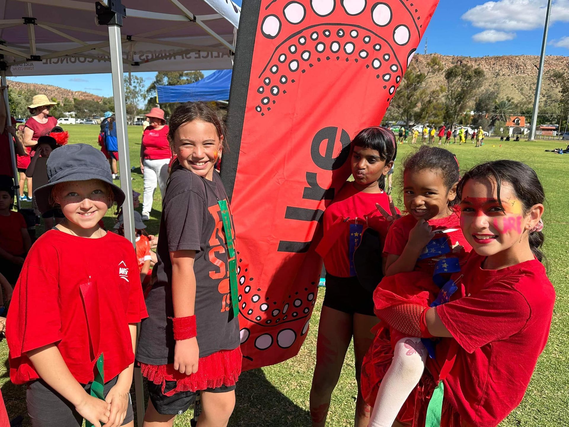 Group of smiling kids in red shirts and face paint at outdoor event.