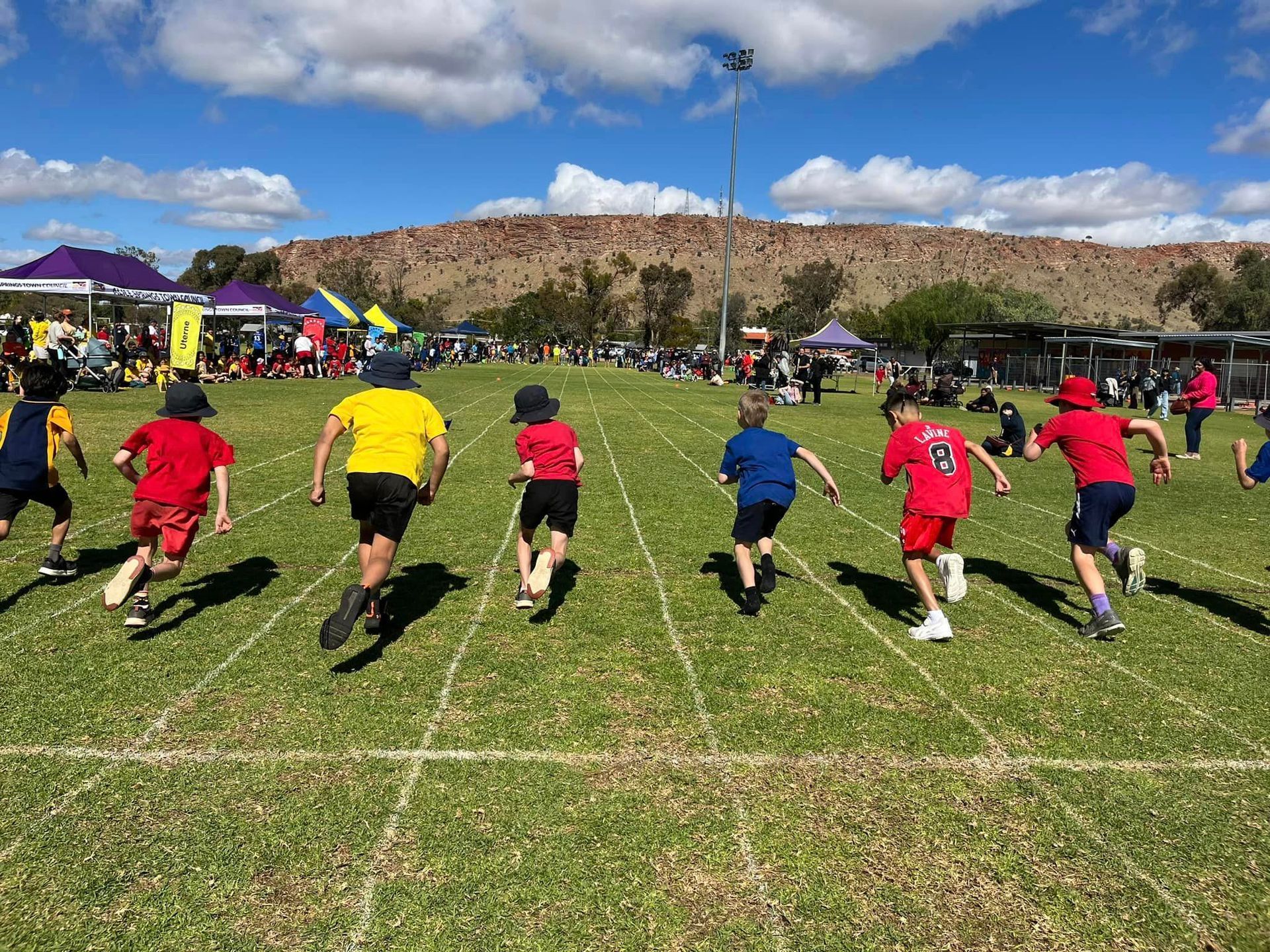 Children racing on a green field; red, yellow, and blue shirts; spectators and a hill in the background.