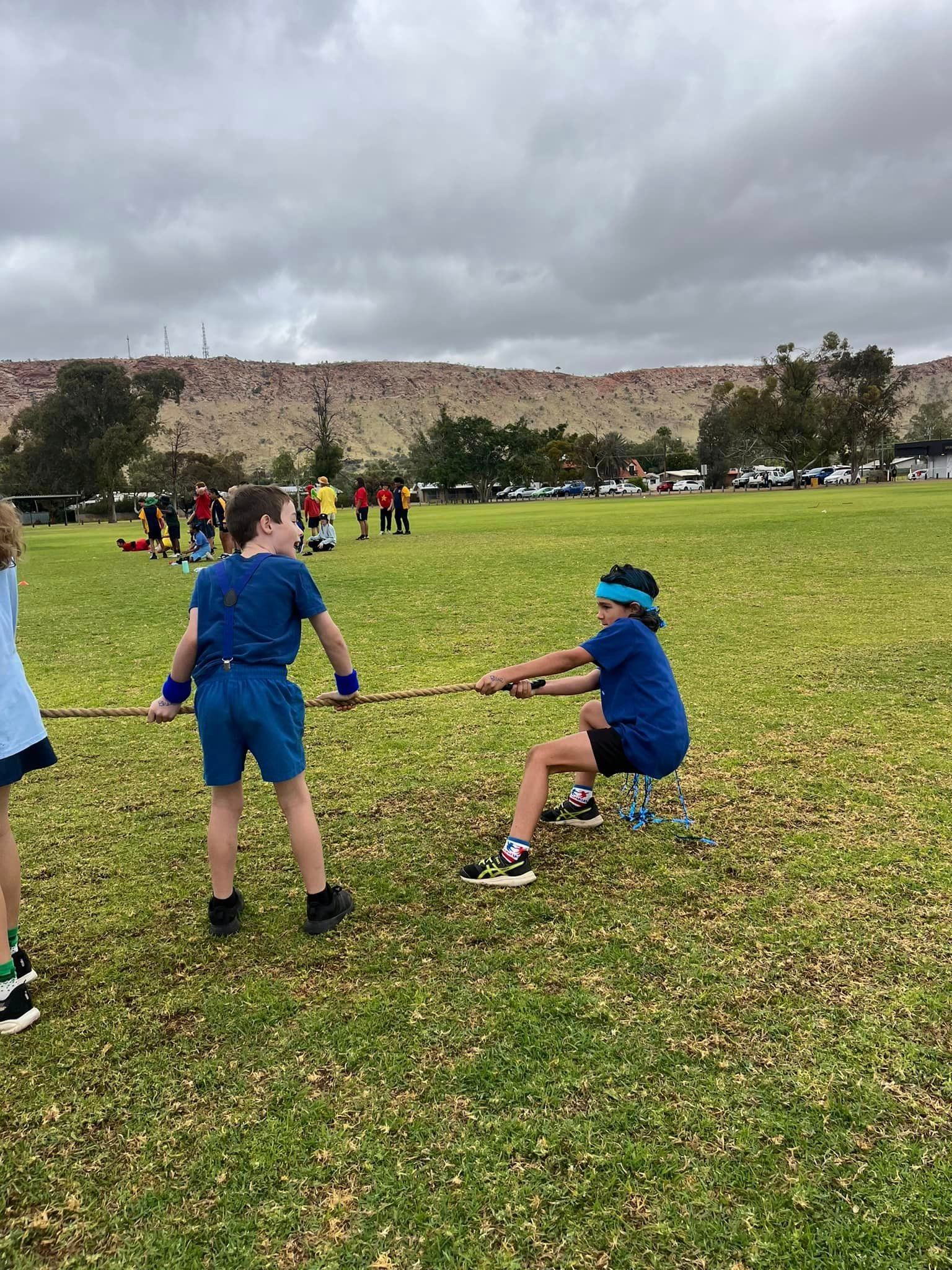 Children playing tug-of-war on a grassy field under a cloudy sky. One child blindfolded.