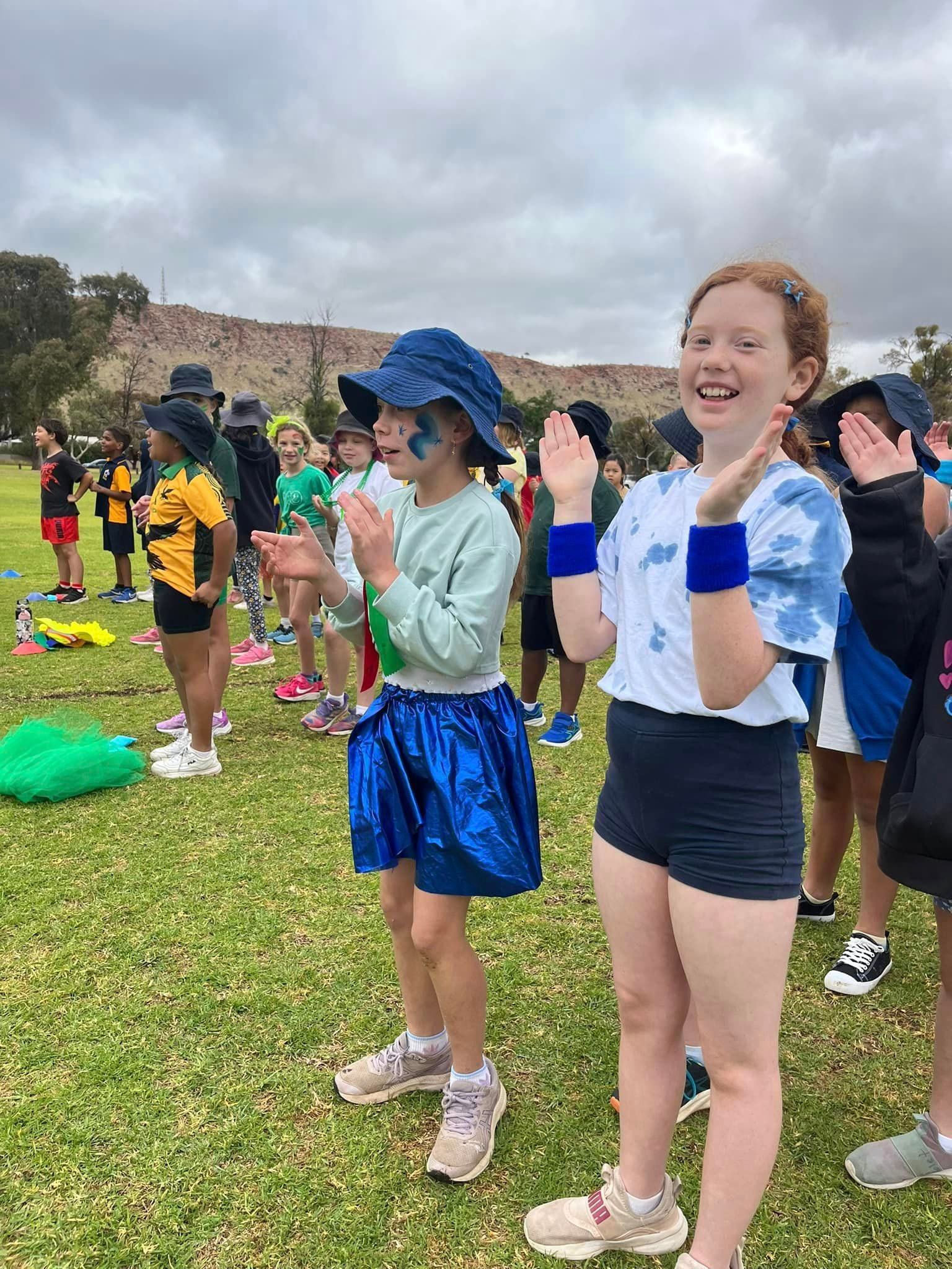 Children in costume clapping on a grassy field with a cloudy sky background.