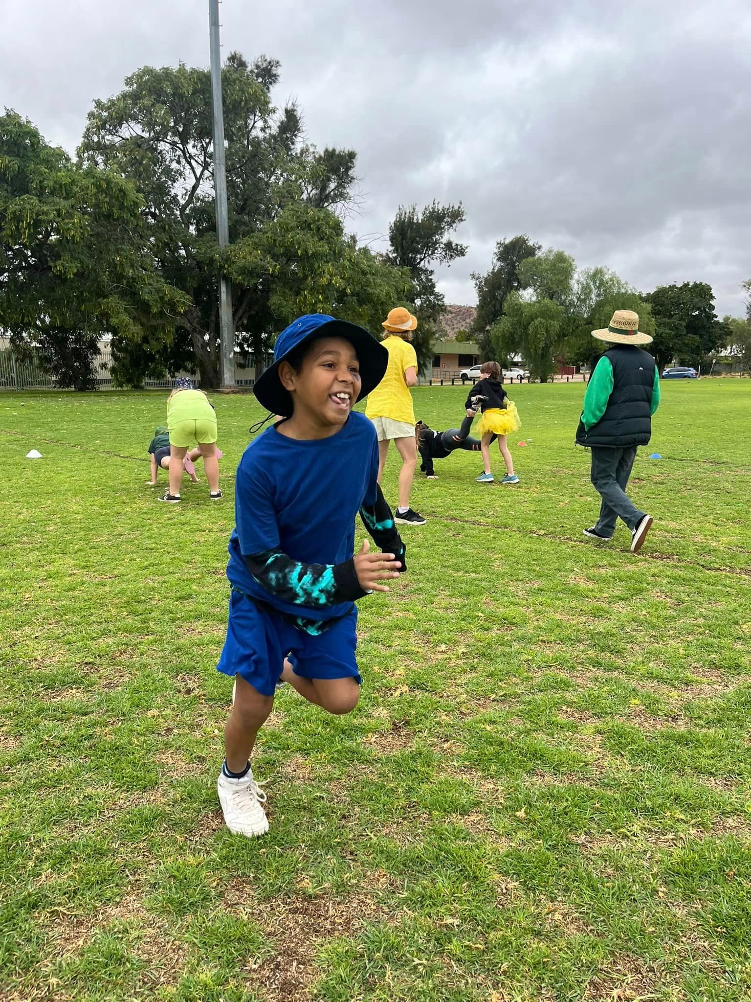 Boy running across a grassy field, smiling. Other children are in the background. Cloudy day.