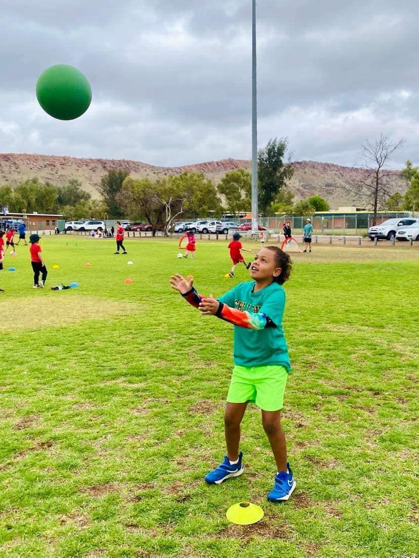 Young child in green catching a green ball on a grassy field, with other children and hills in the background.