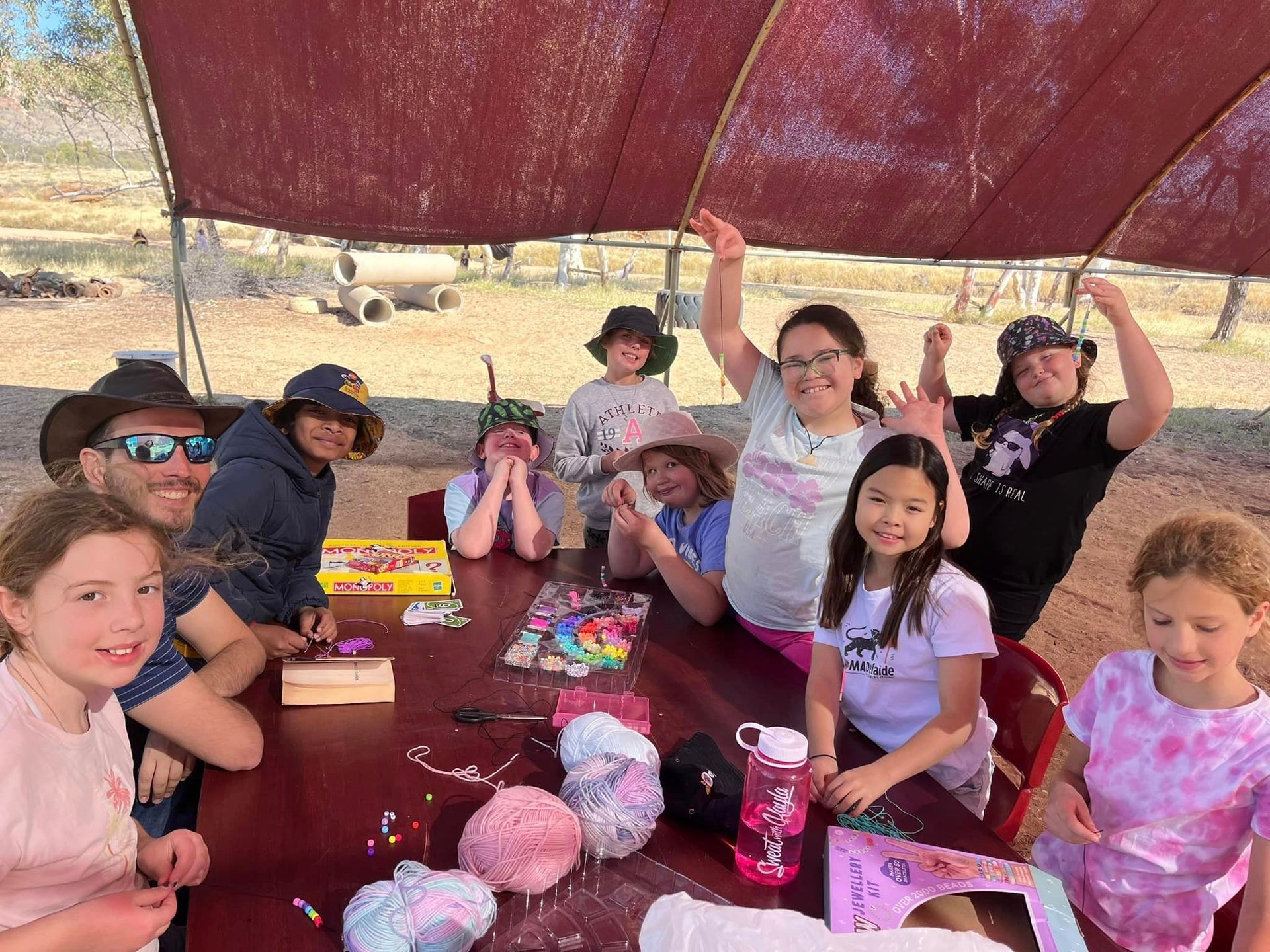 A group of children and adults are gathered at a table under a red awning, crafting and smiling.