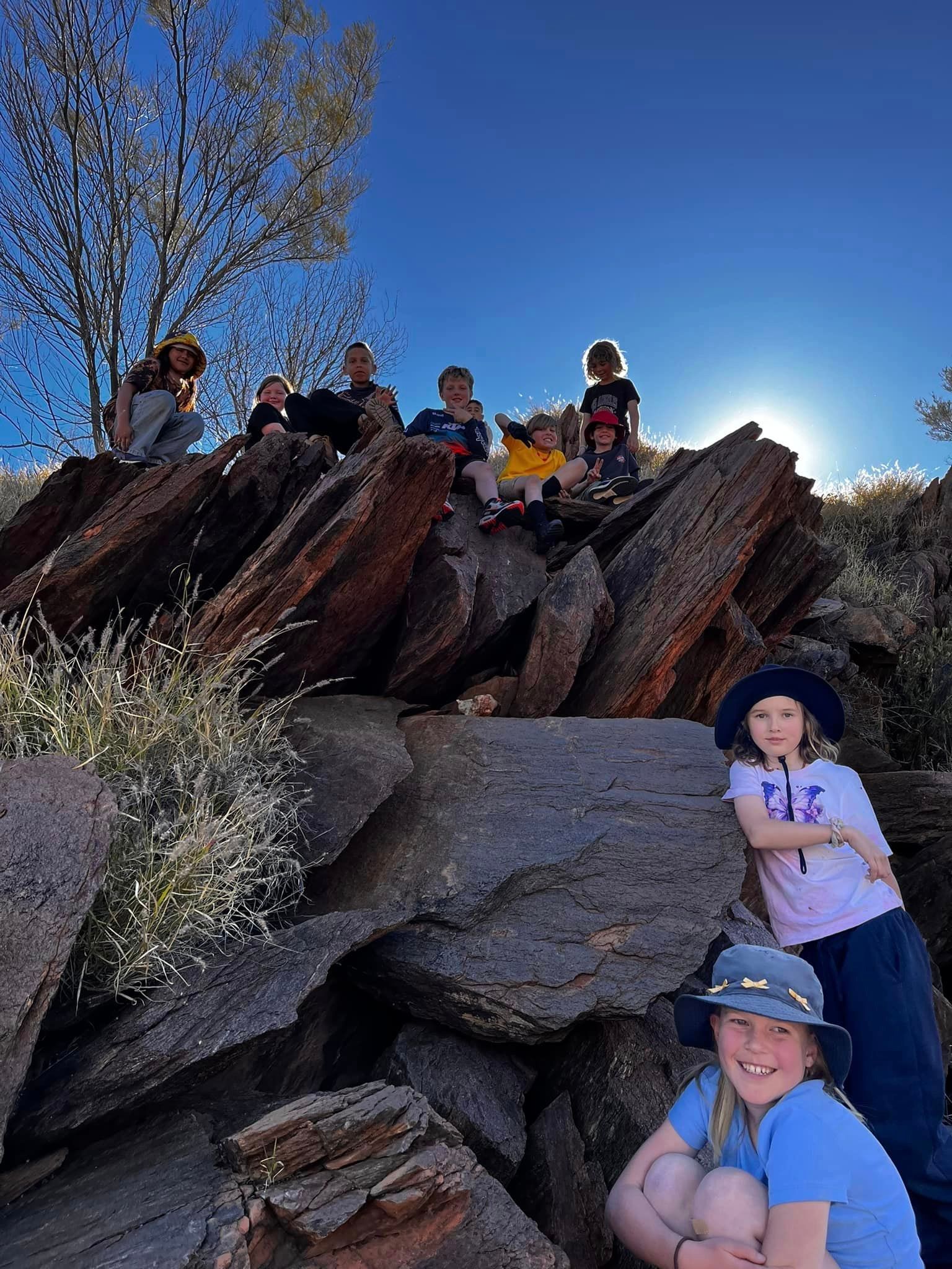 Kids on rocks, blue sky, sun. Group smiles, nature setting, brown rocks, hats.