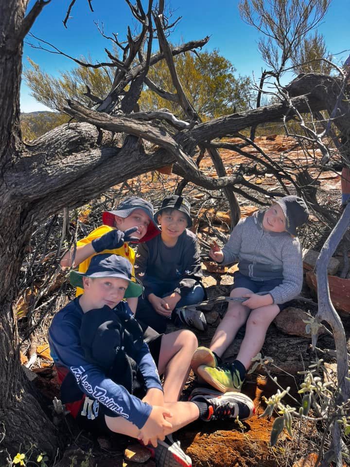 Four children sitting under a tree in a desert landscape, smiling and looking at the camera.