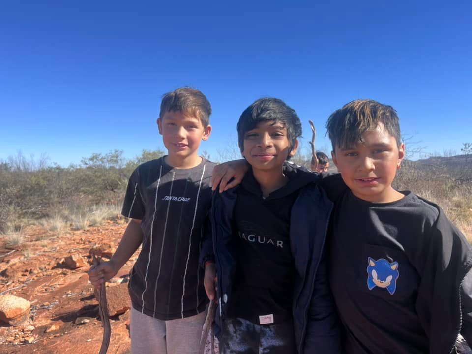 Three boys posing outdoors, smiling at the camera. They're in a dry, sunny environment with red soil.