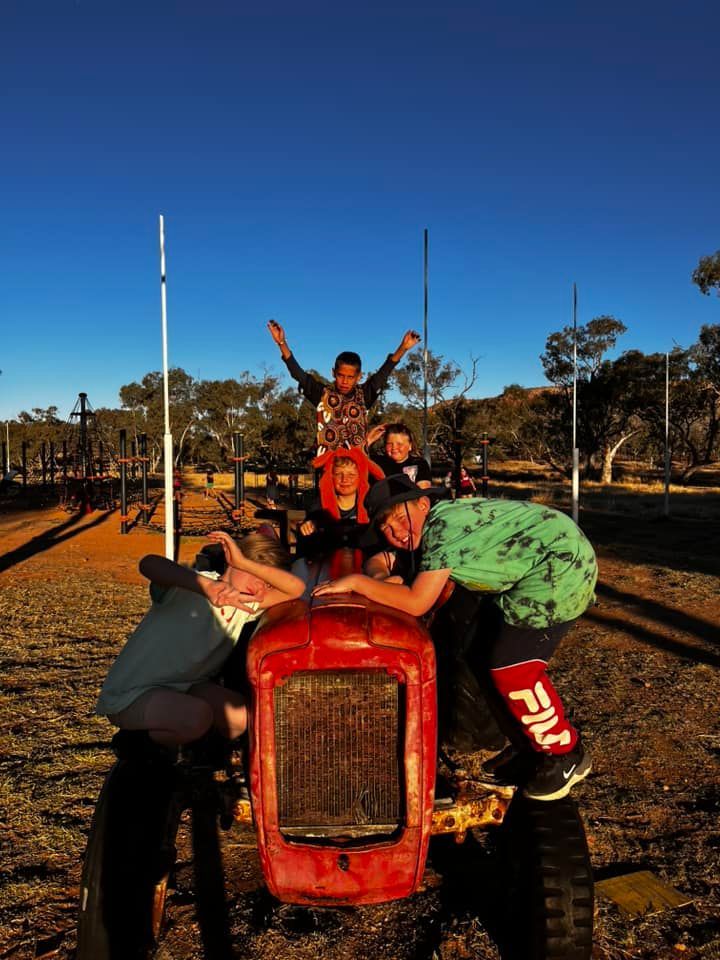 A group of five people posing on a red tractor in a sunny field, arms raised, smiling.